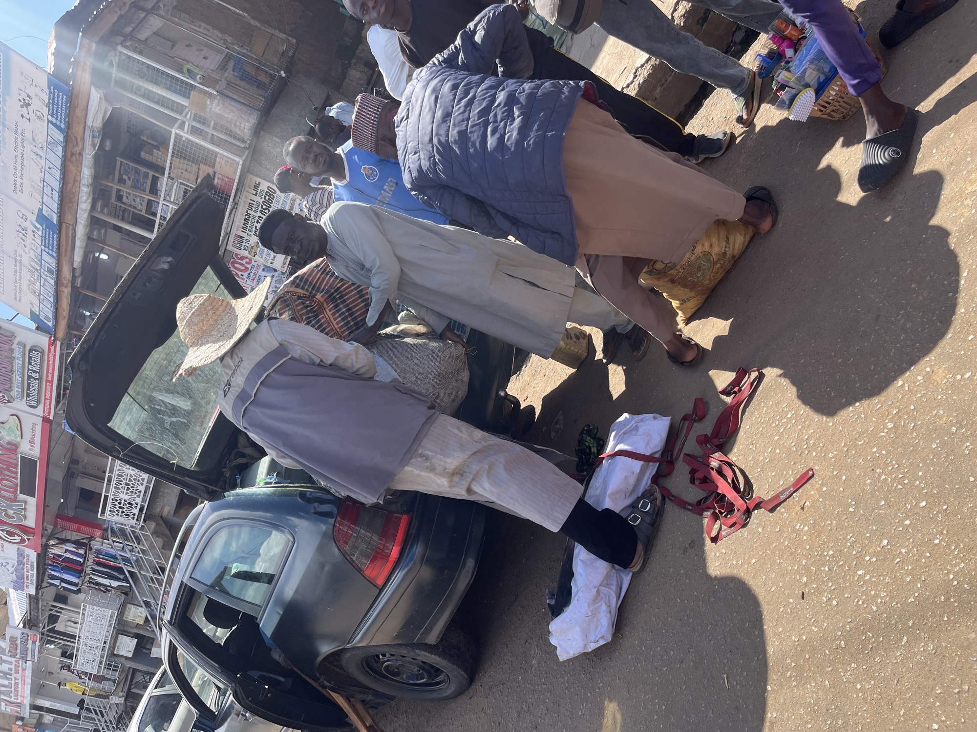 A group of people are gathered around an open car trunk on a street in Jos, Nigeria. A dark-colored station wagon has its trunk open, and several individuals are involved in loading or unloading items. One person in a gray robe and a straw hat appears to be assisting with a large, striped bag. Another person is partially obscured by the car, wearing a blue quilted jacket and a light brown outfit. Red straps are visible on the ground near a white cloth-wrapped object. The surrounding environment suggests an urban setting with buildings and shops lining the street, characterized by signs for clothing and electrical goods. The ground is paved with asphalt. The lighting indicates it is daytime.