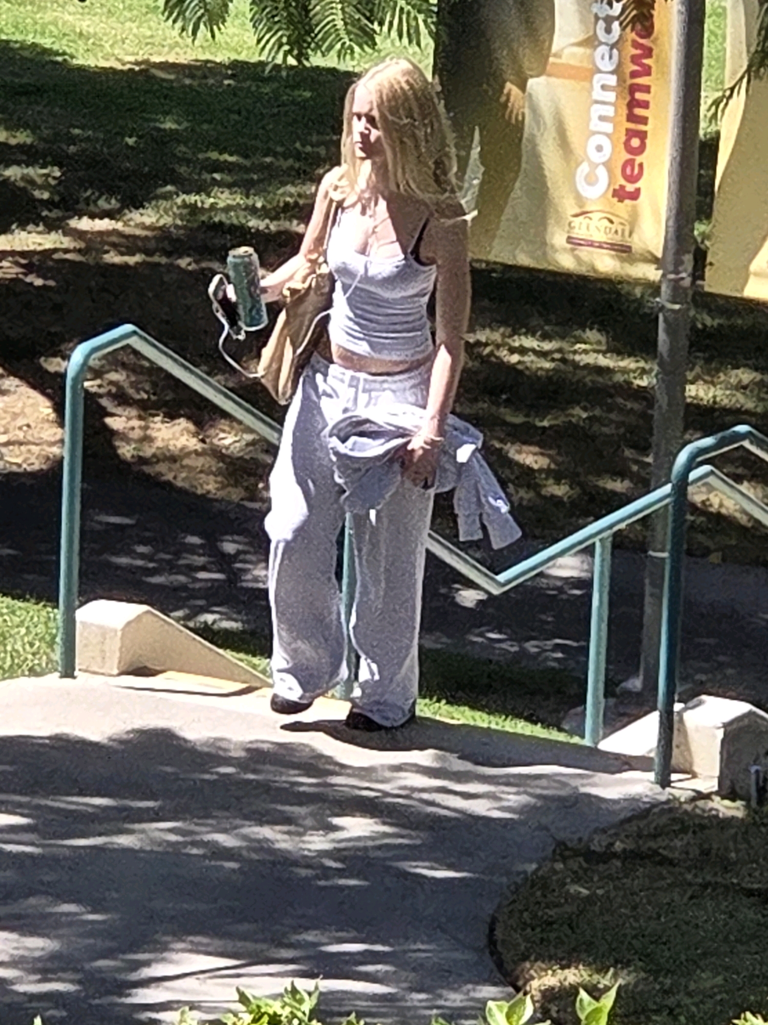 In Glendale, California, a Caucasian female with long, blonde hair walks down a paved walkway with metal handrails. She is wearing a white tank top, grey sweatpants, and dark shoes. In her right hand, she holds a can and a phone with earbuds. A light-brown tote bag hangs over her left shoulder. The background consists of trees and a banner that reads "Connect teamwork" and "Glendale." The walkway is illuminated by dappled sunlight, casting shadows on the ground.