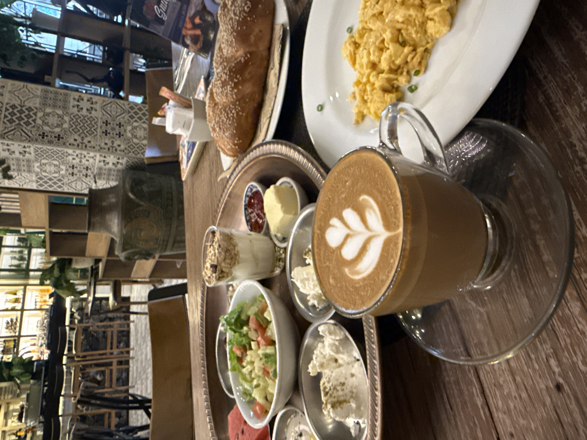 A close-up, high-angle shot captures a breakfast spread on a wooden table, likely in a restaurant in Ko Samui District, Thailand. In the foreground, a clear glass mug of coffee with latte art rests on a matching saucer. To the right, a white plate holds a serving of bright yellow scrambled eggs, garnished with a few green chives. In the middle ground, a large, oval-shaped metal tray holds various breakfast items. This includes a bowl of salad with tomatoes and cucumbers, a glass of yogurt with granola, a small dish of jam, a cube of butter, and two small bowls containing white cheese, one sprinkled with herbs. To the right of the tray, a large, golden-brown loaf of bread, covered in sesame seeds, sits on a white plate. In the background, the scene opens up to a blurred view of a restaurant interior, featuring a bar area with stools, shelves stocked with bottles and decorative items, and patterned tilework on a wall. The overall impression is a satisfying breakfast meal at a well-appointed establishment.