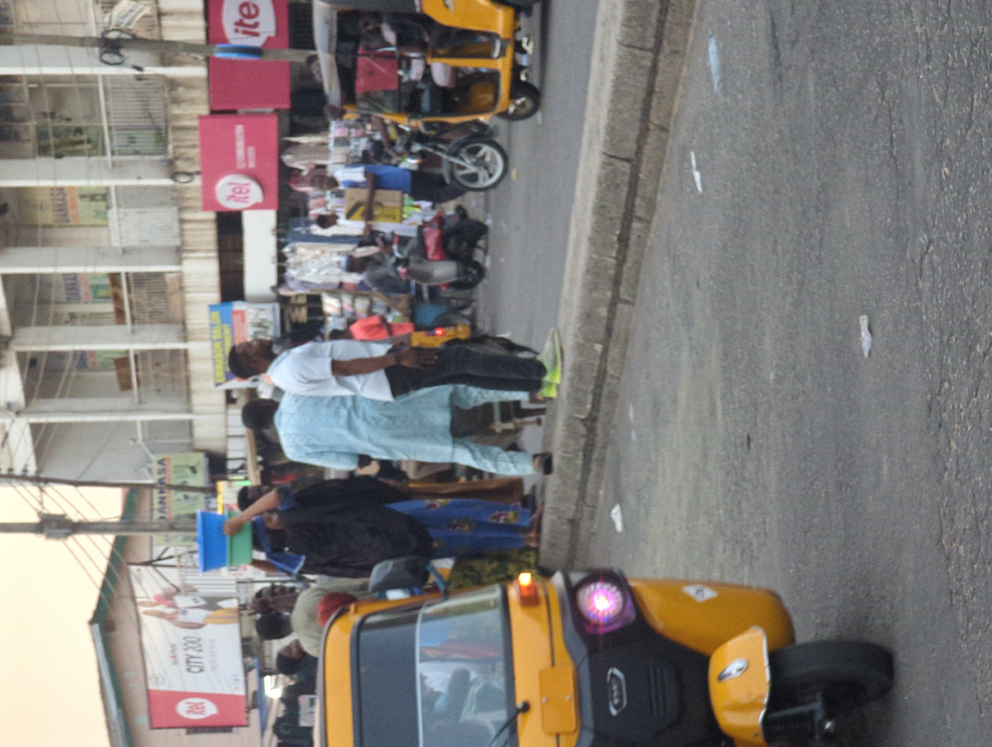 The image depicts a street scene in Maiburiji, Nigeria. People are walking on the sidewalk next to a street with yellow auto rickshaws (tuk-tuks). There are buildings in the background with advertisements for "itel". Some people are carrying boxes. The street is paved and appears to be somewhat busy with vehicular and pedestrian traffic.