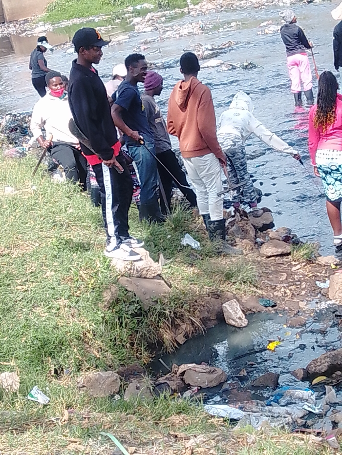 A group of people stands along a polluted riverbank and in the river itself, possibly cleaning it. Most hold tools like rakes or sticks. The river is filled with garbage and debris, reflecting the light. A separate, smaller polluted stream runs off from the main river in the foreground. The riverbank is covered with grass and scattered rocks.
