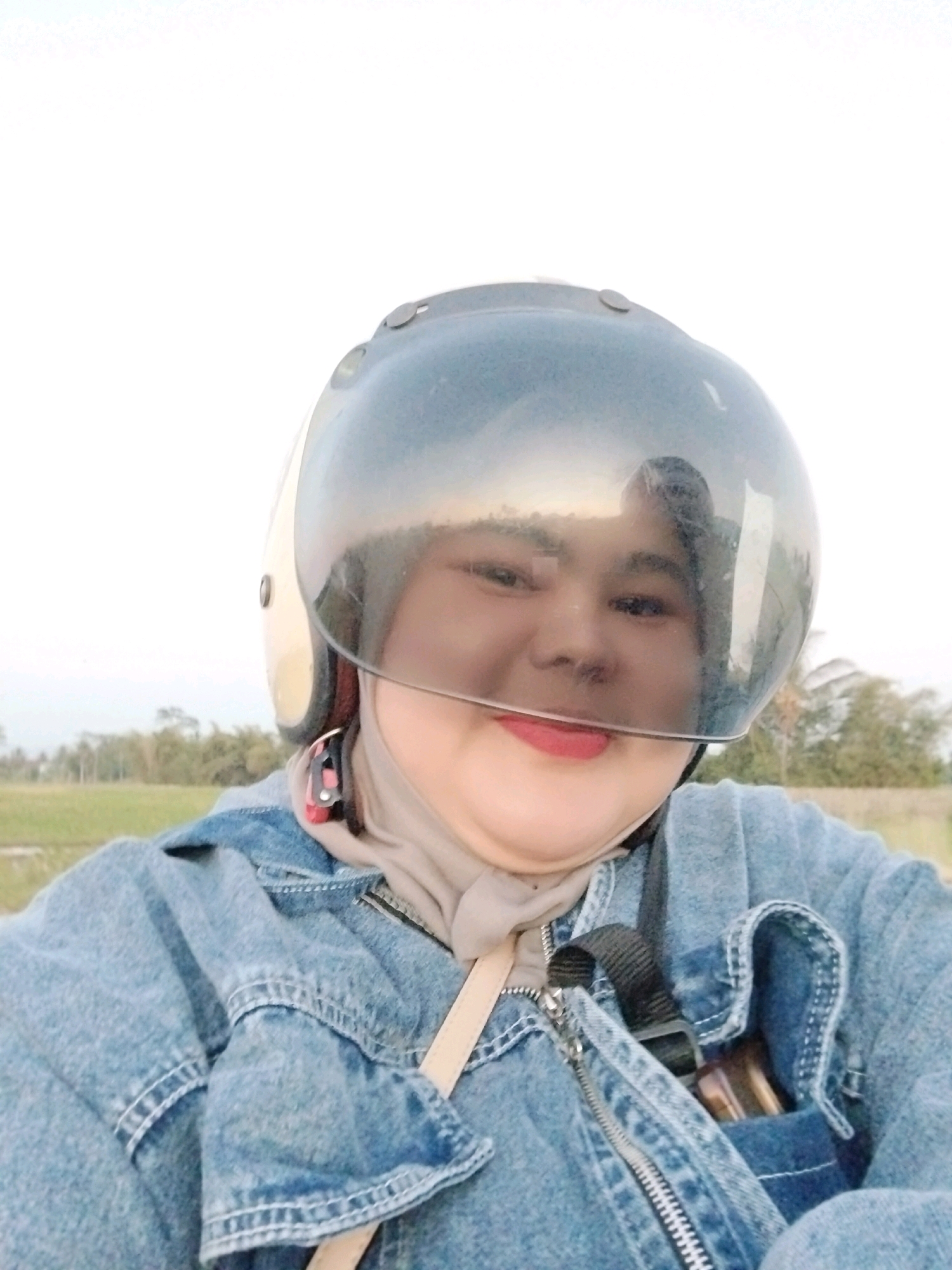 A close-up, selfie-style medium shot shows a young woman wearing a light beige hijab and a denim jacket. She is also wearing a white and gold motorcycle helmet with a clear visor that reflects the sky and trees. Her face is visible through the visor, and she is looking towards the camera with a faint smile. A strap from a light-colored bag is visible across her chest. The background consists of a bright, overcast sky and a blurry landscape of trees and what appears to be a field, suggesting an outdoor setting in a rural or suburban area, potentially Linggasirna, Indonesia. The lighting is soft and diffused, typical of an overcast day.