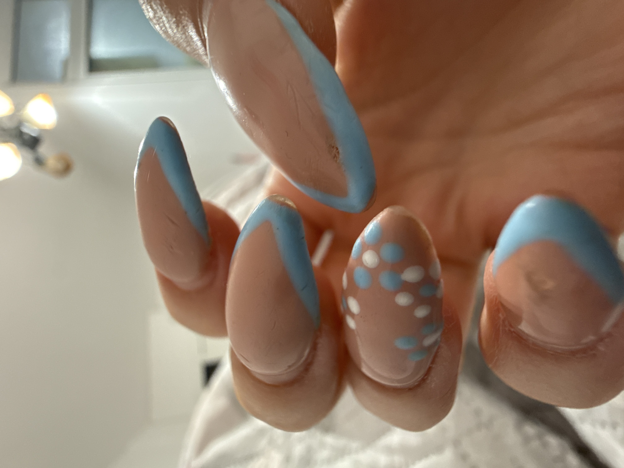 A close-up view of a hand displaying painted fingernails with a blue-and-nude color scheme. The nails are long and almond-shaped. One nail features a pattern of blue and white polka dots. The background is blurred.
