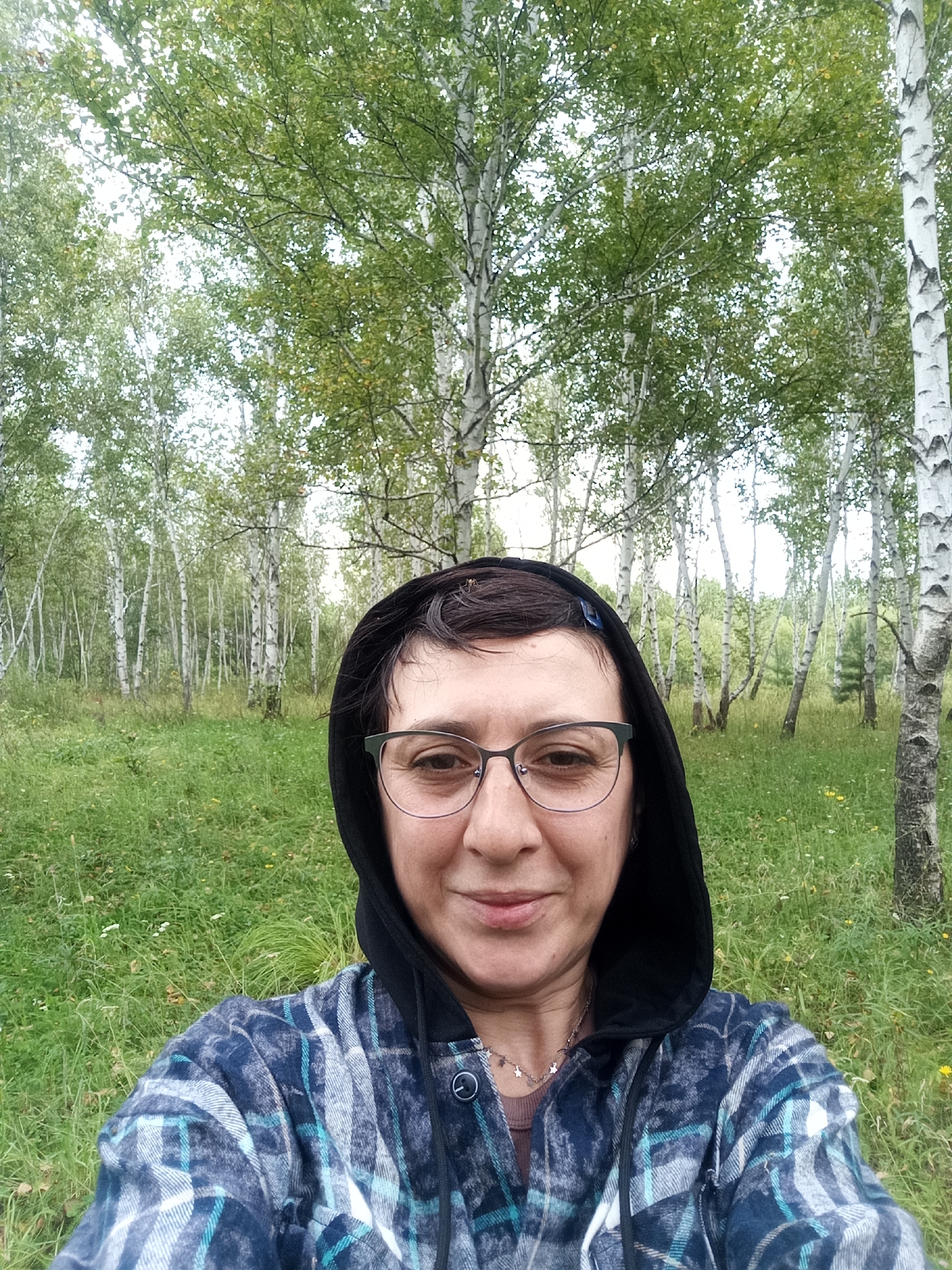 A selfie is taken by a person with short, dark hair and glasses, wearing a dark hooded jacket over a plaid shirt and a star-patterned necklace. The person is smiling faintly, with their face turned towards the camera. The background features a dense forest of birch trees with white bark and green foliage, interspersed with green grass and some scattered wildflowers. The scene appears to be an outdoor setting, likely in or around Nikolskoye, Russia, given the prevalence of birch forests in the region. The lighting suggests an overcast day.