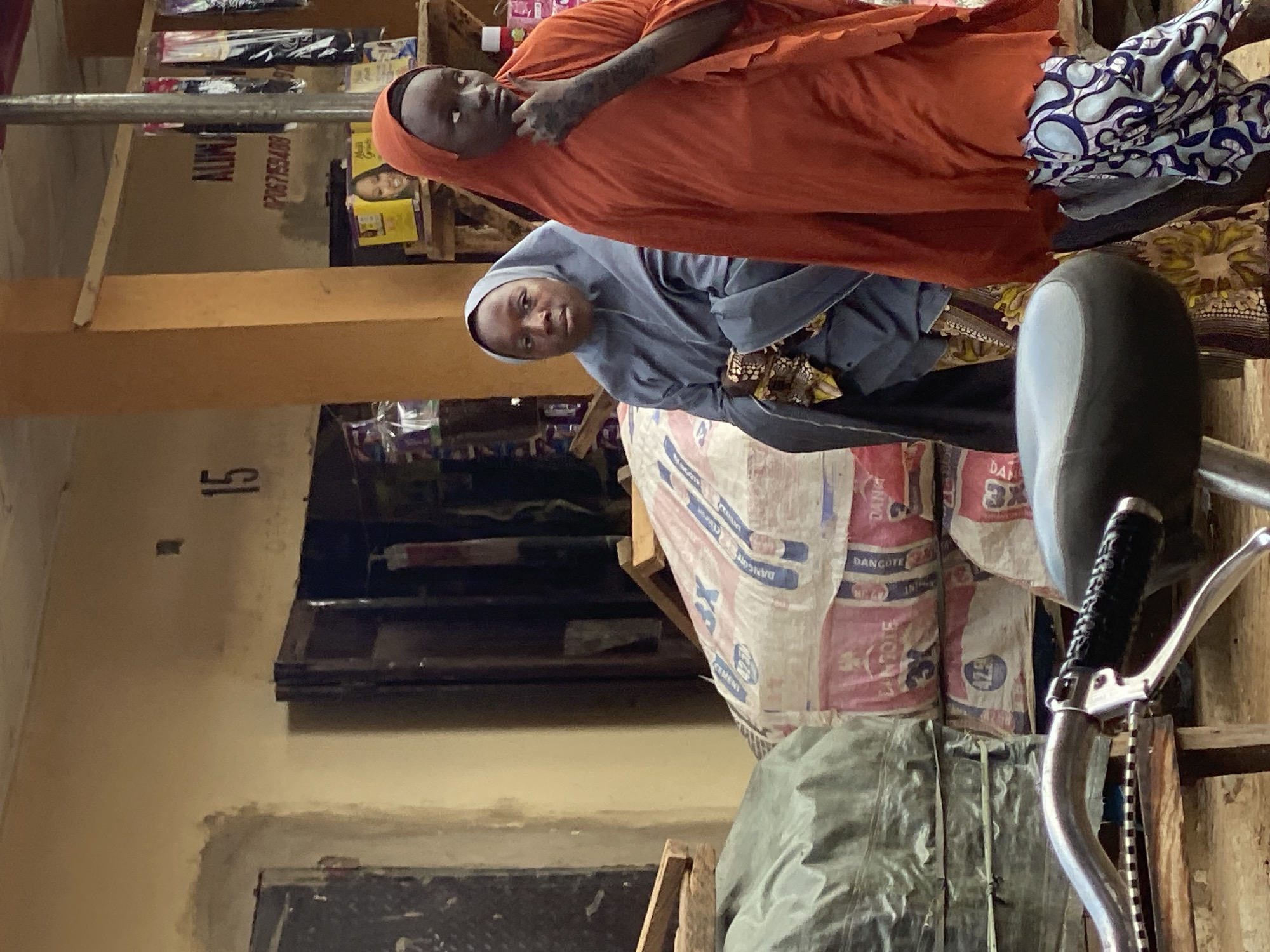 The image, taken in Kafanchan B, Nigeria, shows two women in a building. One woman is wearing an orange hijab and dress, while the other wears a grey hijab and dress. Both are looking towards the camera. The background features bags of Dangote cement stacked up. Also in the background, the number 15 can be seen on a wall. A bicycle handlebar and seat are in the foreground. Merchandise is visible on shelves in the background.