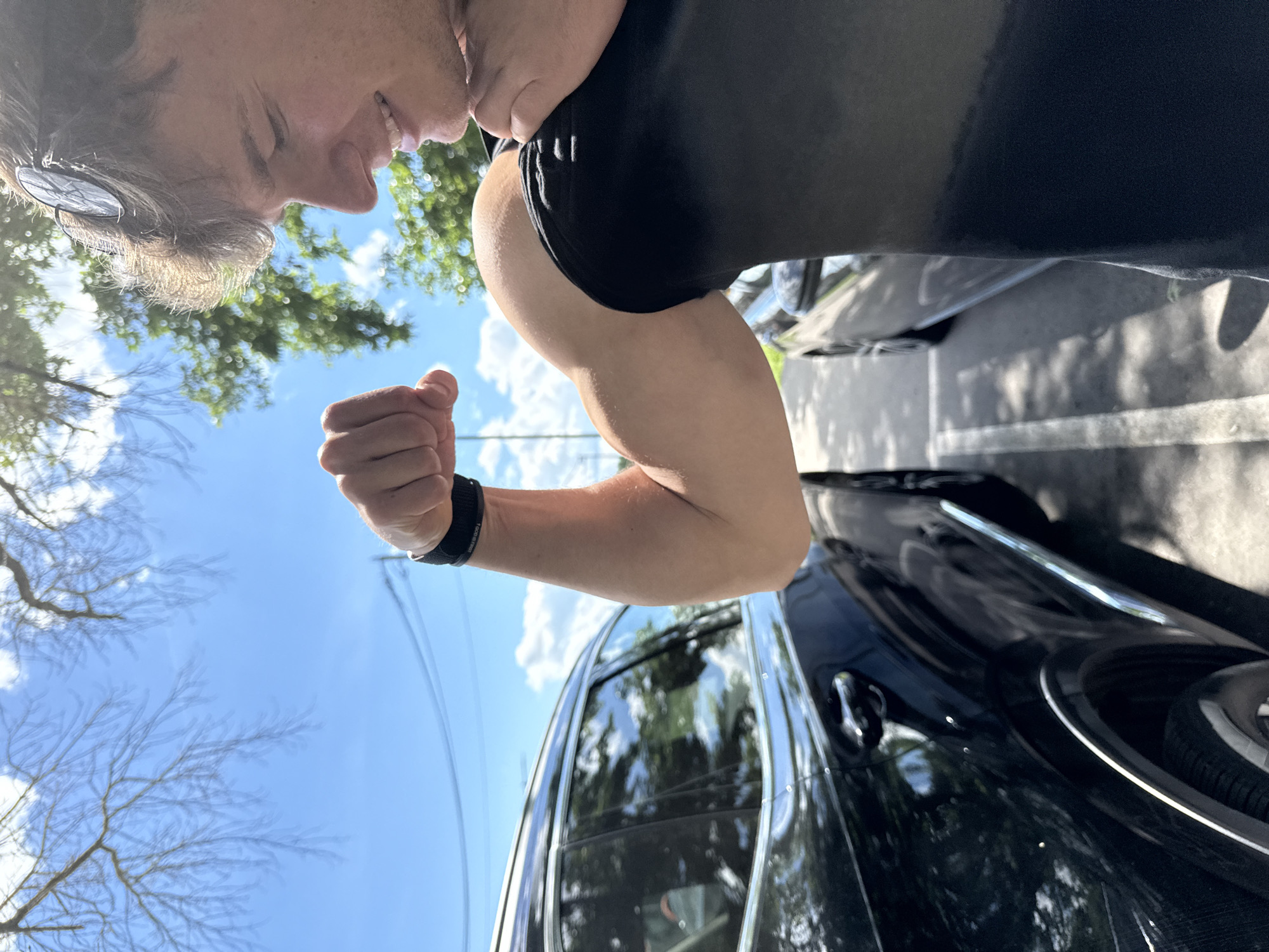 The image depicts a person flexing their right arm, displaying their bicep muscle. The person is wearing a black athletic shirt and a black fitness tracker on their wrist. The background features a black car and trees set against a bright blue sky with scattered clouds. The location appears to be outdoors, likely in an urban or suburban environment with parked vehicles. No specific information confirms this is Elhovo, Bulgaria.
