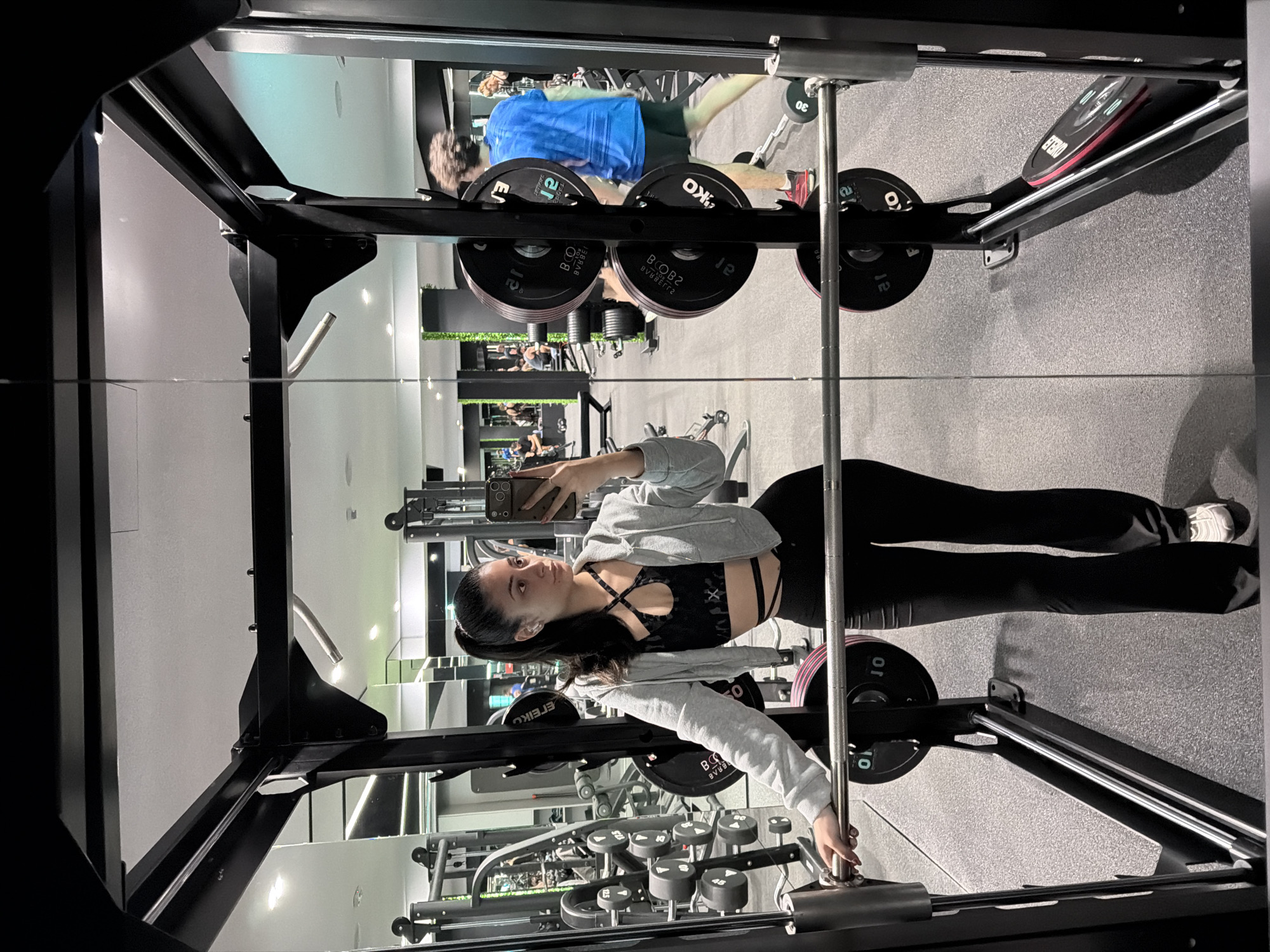 Here is a forensic description of the image:
The image shows a gym interior, likely a fitness center, with a woman taking a selfie using her smartphone. She is positioned in front of a mirrored surface within the gym equipment, providing a dual view of the scene. The woman is dressed in a black sports bra, black leggings, and a grey hoodie. She is holding her phone up to capture the photo.
In the background, there is gym equipment, including weight racks, dumbbells, and other exercise machines. Another person, a man, is seen on the opposite side of the room, engaged in a workout. The floor appears to be a grey, rubberized material. The environment is brightly lit with modern lighting fixtures.
The precise location is impossible to determine from the image.