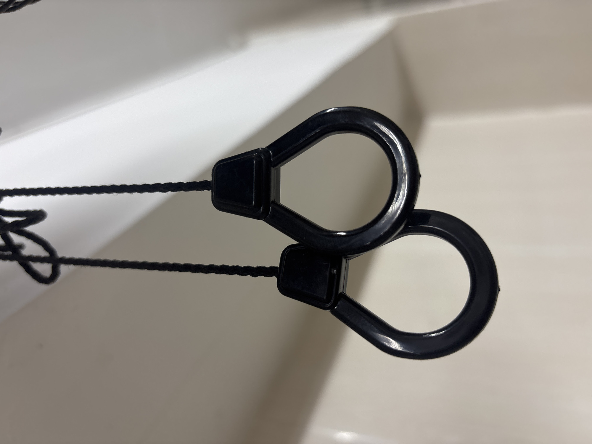 The image shows two black plastic ring handles suspended from black braided cords. The handles are teardrop-shaped, with the wider part of the teardrop forming the ring that one would grasp. The cords are attached to the top, narrower part of the handles. The background appears to be a plain, light-colored wall with some shadows and highlights, suggesting an indoor environment. There are no people or discernible urban elements in the image. The location is Unknown, Unknown.
