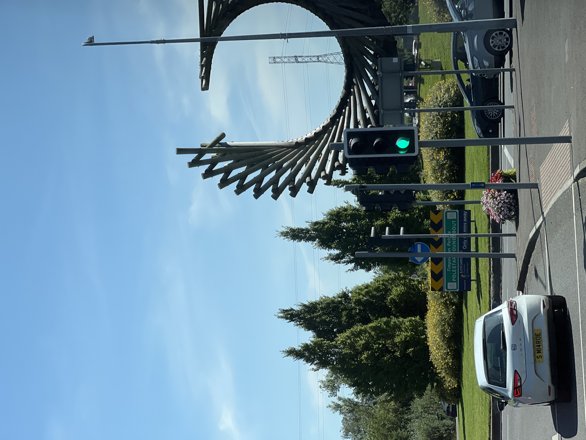 This is a photograph taken from a vehicle at a traffic light, facing a large roundabout with a prominent spiral sculpture made of wooden poles. The sculpture is positioned in the center of the roundabout, which is surrounded by greenery and flowering bushes. To the left of the sculpture, there are several tall, conical evergreen trees. A street light with a camera attached is visible to the right of the sculpture.

In the foreground, a white car is stopped at a red traffic light. The car has a license plate that reads "S MI4ROE". Two other cars are visible on the road beyond the white car, one black and one gray. A traffic sign indicating "Timberlagh, Polestar Roundabout" is visible behind the white car.

The sky is clear and blue with a few wispy clouds. The overall scene appears to be a sunny day in an urban or suburban setting. The location is identified as Polestar Roundabout in Letterkenny, Ireland.