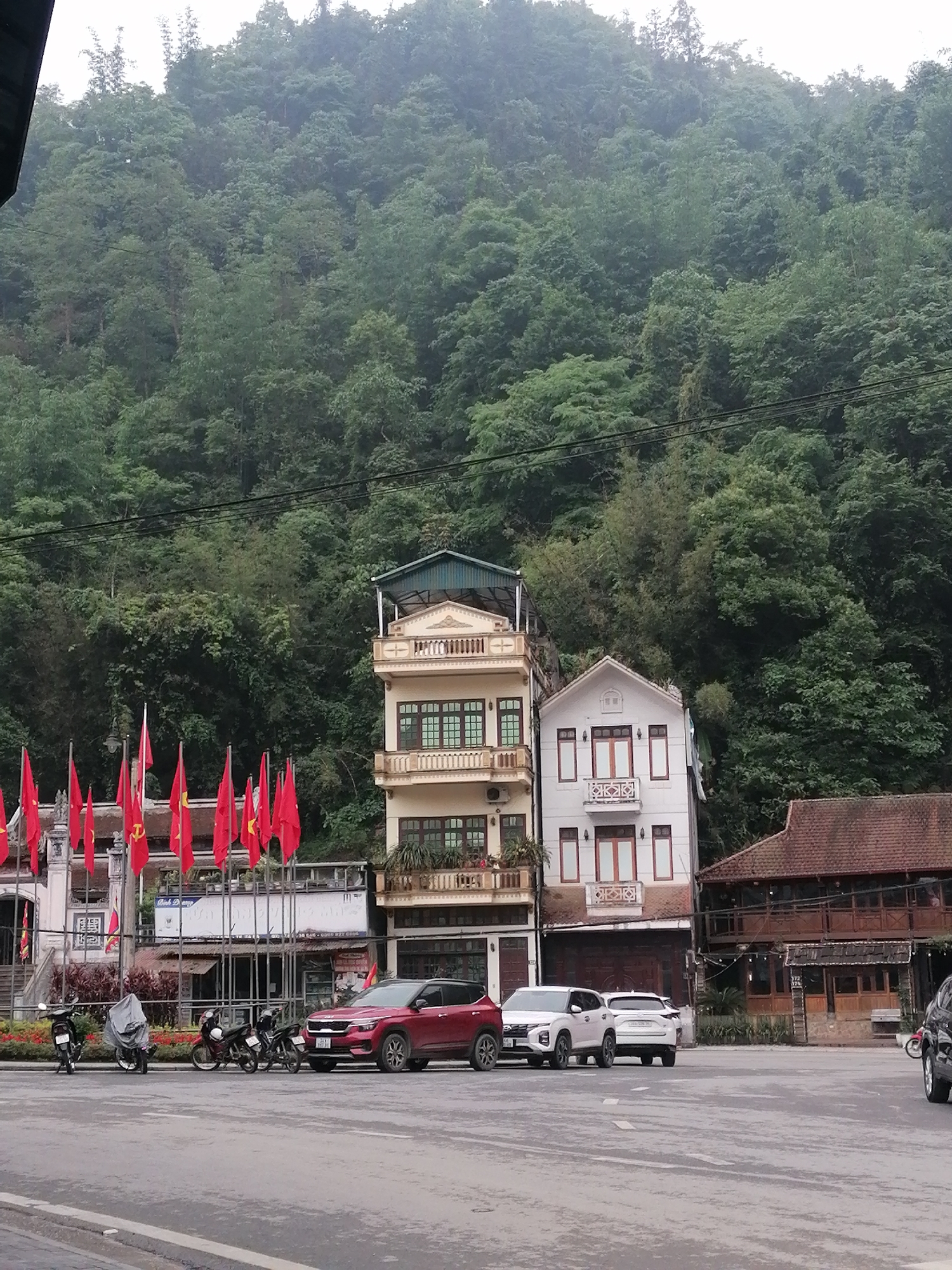 The image depicts a street scene in Sa Pa Ward, Vietnam. In the foreground is an asphalt street with parked vehicles, including a red SUV, two white SUVs, and several motorbikes. Red flags are prominently displayed along the side of a building, possibly signifying a political or national event.
Behind the vehicles, there are buildings with distinct architectural styles. One building is a three-story structure with yellow facade and balconies, while another is a white building with a gable roof and minimal ornamentation. Adjacent to these is a wooden structure with a traditional Southeast Asian design.
The background is dominated by a lush, green hillside covered in dense vegetation, characteristic of the mountainous terrain in the region. Overhead, power lines run across the frame. The scene appears to be captured during daylight hours under overcast lighting conditions.