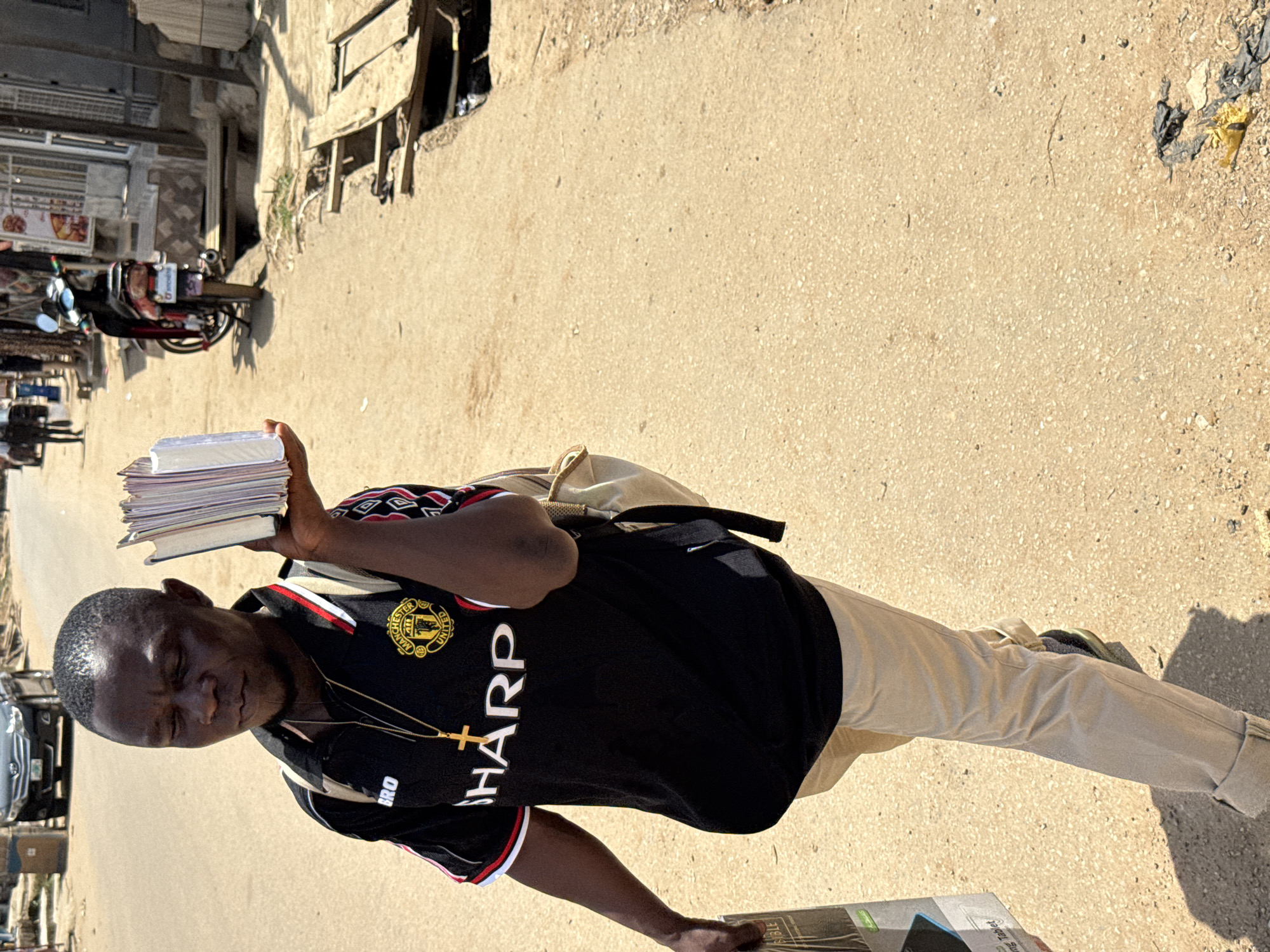 A young Black man is standing on a dirt road under bright sunlight. He is holding a stack of books in his left hand and his right arm is bent at the elbow, resting his hand on his chest. He is wearing a black Manchester United jersey with the team's crest on the left breast and a gold cross necklace. His pants are a light tan color, and they are cuffed at the bottom. He appears to be looking down with his eyes closed or looking away from the camera, with a serene expression on his face. Behind him, there is a motorcycle parked on the side of the road, and in the background, there are some buildings and structures. The scene suggests a warm, sunny day, likely in a semi-urban or rural area in Abuja, Nigeria. The overall impression is one of a person in quiet contemplation or perhaps taking a moment of rest. Visible text includes "MANCHESTER UNITED" on his jersey and "SHARP" below it.