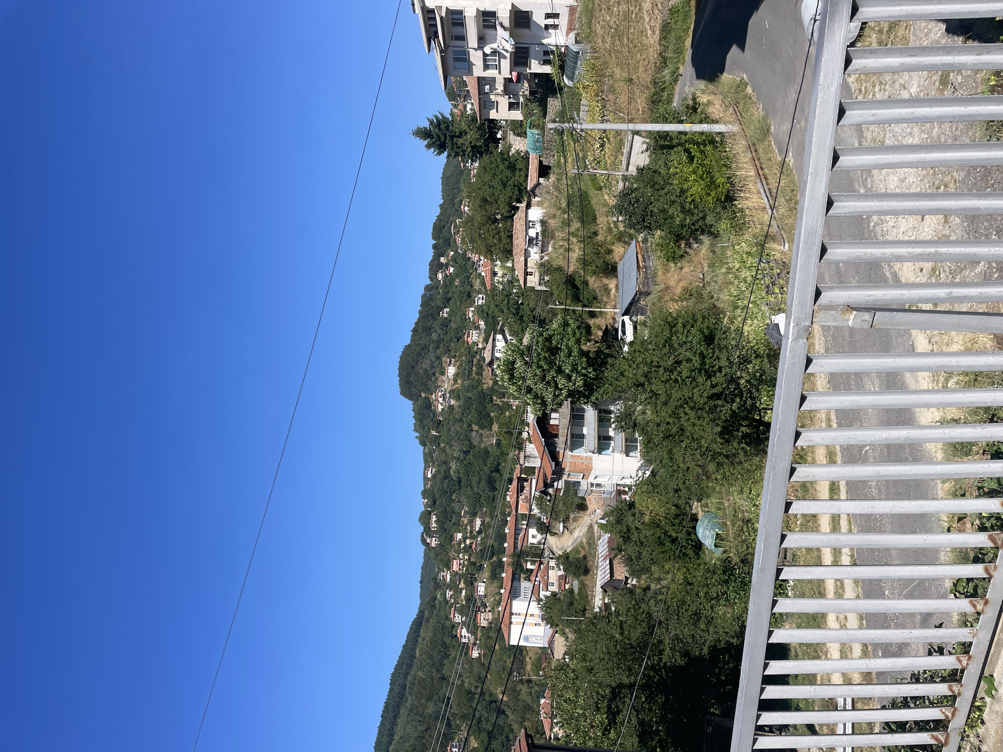 The image is a medium shot of a village nestled in a hillside. The sky is a clear, bright blue, with a thin power line stretching across it. The village is comprised of numerous houses with red-tiled roofs, scattered across the green, tree-covered slopes. In the foreground, there is a metal railing. The houses vary in size and architectural style, with some appearing to be older stone structures and others more modern buildings. Lush green trees and vegetation cover the landscape, indicating a natural, rural environment. The overall impression is one of a peaceful, traditional village situated in a hilly, scenic area. The village is confirmed to be Davidkovo, Bulgaria.