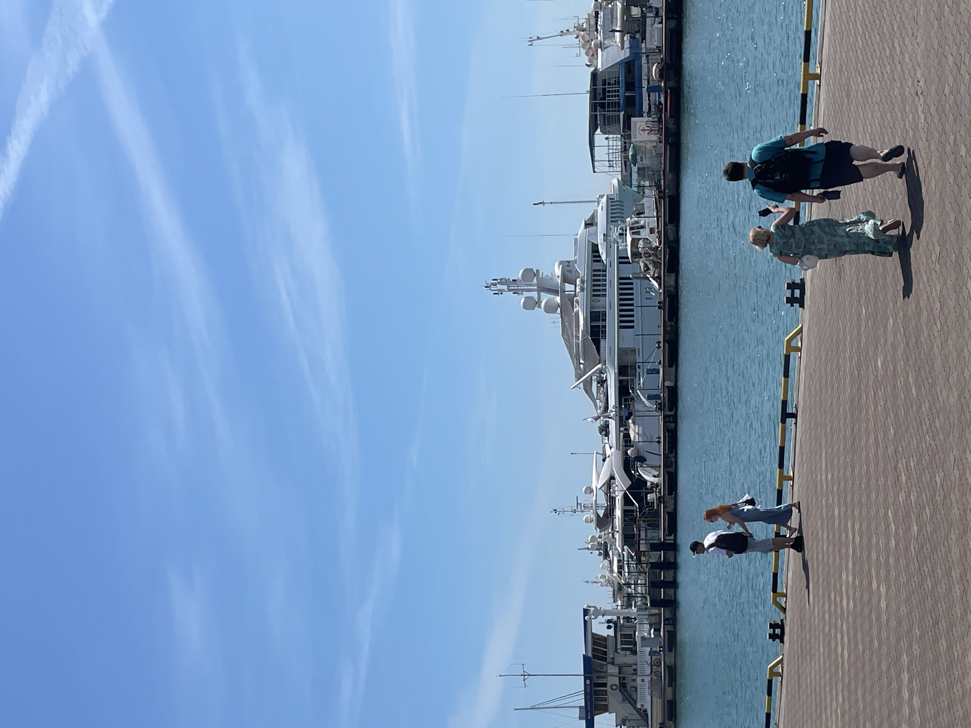 This image depicts a sunny daytime scene at the Sochi Commercial Sea Port in Sochi, Russia. The foreground features a reddish-brown paver-tiled promenade, bordered on the right by a yellow and black striped safety railing and multiple black metal bollards. Two pairs of individuals are walking on the promenade.
On the left, a man wearing a white t-shirt, light shorts, a dark baseball cap, and a black backpack walks alongside a woman in a long, light-colored patterned dress with a white shoulder bag and reddish-brown hair. Their shadows are cast on the pavers to their right.
Further to the right and slightly closer to the viewer, a second pair walks away from the camera. A woman in a long, greenish-blue patterned dress, with light hair and a white handbag, holds a smartphone in her right hand. She is walking next to a man wearing a blue t-shirt, dark shorts, a black backpack, and holding a dark object in his left hand. Their shadows are visible on the promenade.
Beyond the promenade is a body of calm blue-green water, reflecting the sunlight. Docked along the far side of the water are numerous vessels. The prominent features include several large, multi-deck luxury yachts, primarily white, equipped with multiple radar domes, antennas, and navigation equipment. Other vessels, including smaller boats and what appear to be utilitarian or passenger craft, are also present, some with blue and white hulls. Structures on the docks are partially visible behind the boats.
The sky occupies the upper portion of the image and is a clear, bright blue with several faint, linear white contrails from aircraft visible across different sections.