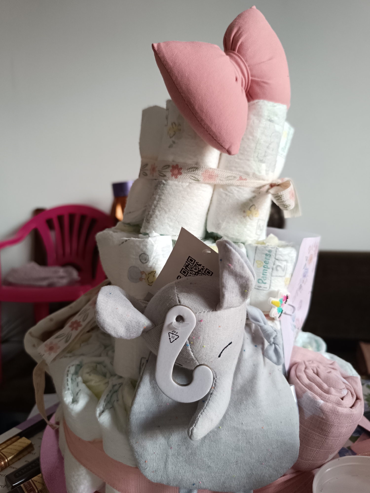 The image shows a diaper cake, a multi-tiered structure made of diapers, decorated with a pink bow on top, a ribbon with floral patterns around each tier, and a stuffed elephant toy hanging from the side. The diapers are white with cartoon prints. The background is a blurry mix of a pink plastic chair, a white wall, and other indistinct objects, indicating an indoor setting, possibly a home or a baby shower venue.