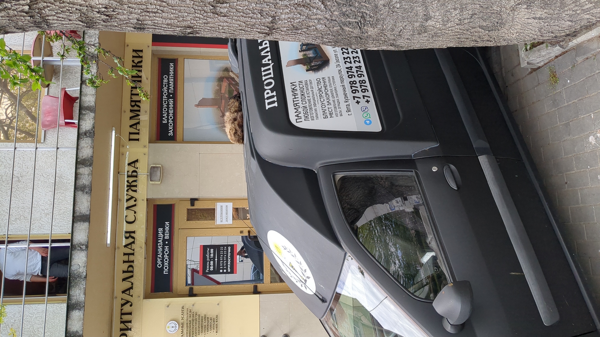 A matte black van, parked on a sidewalk next to a gray tree trunk, displays signage with the word "ПРОЩАЛЬ" (farewell) and contact information. It is parked outside a "РИТУАЛЬНАЯ СЛУЖБА" (funeral service) and "ПАМЯТНИКИ" (monuments) establishment. The address, displayed on the sign, is город Ялта, Кузнечный переулок, 2А (Yalta, Kuznechny lane, 2A). Through the shop window, a person is visible in a white shirt.
