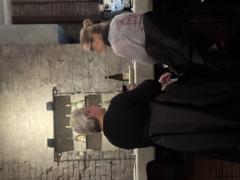 Two individuals are seated at a counter within a dimly lit restaurant or bar in New York, United States of America. The background features an exposed light-colored brick wall with a recessed shelf display containing several bottles. A middle-aged male with grey hair, wearing a dark long-sleeved shirt, is seated on the left, facing right. He holds a glass containing red liquid, likely wine. His left forearm displays a tattoo. Opposite him, a middle-aged female with blonde hair tied in a bun, wearing blue-rimmed glasses and a light-colored long-sleeved shirt with a red lobster graphic on the back, is seated, facing left towards the male. A dark coat is draped over the back of each person's stool. On the light-colored counter between them, a green wine bottle labeled "SANGUE DI GIUDA" and a white dish are visible. To the far right, a coffee machine or similar stainless steel beverage appliance is present.