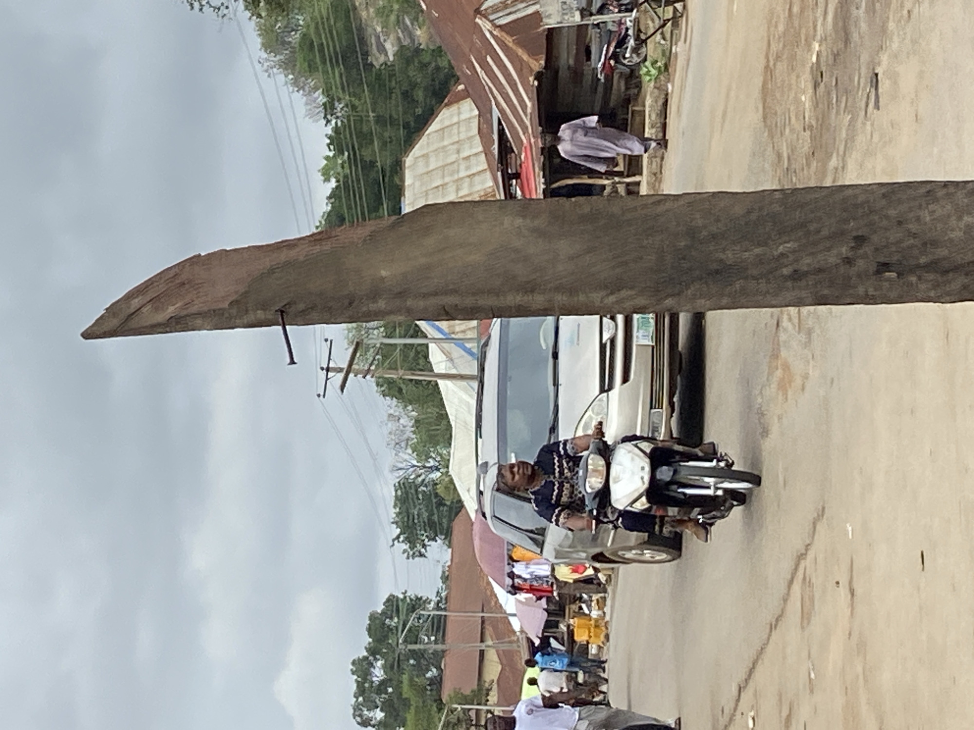 This image captures a busy street scene in Mararaba, Nigeria, under an overcast sky. The photograph is oriented 90 degrees clockwise.
In the foreground, a man wearing a dark blue and white patterned top is riding a motorcycle, looking towards the camera with a smile. Behind him, a white van is parked or moving slowly, with its license plate partially visible as "63 E...". The street appears to be paved but shows signs of wear.
A large, rough-hewn wooden beam extends prominently across the mid-section of the frame, horizontally traversing the street. Below and behind this beam, and adjacent to the white van, are several makeshift stalls, possibly selling clothing or other goods, indicated by colorful fabrics and umbrellas. Other pedestrians are visible walking or standing further down the street, including one person in a white garment.
The background features simple structures with corrugated metal roofs, typical of local architecture. Utility poles with multiple power lines are visible against the uniformly gray, overcast sky. Lush green trees can be seen in the distance, indicating a verdant environment. The overall scene depicts a regular daytime street activity, likely involving commerce and transportation.