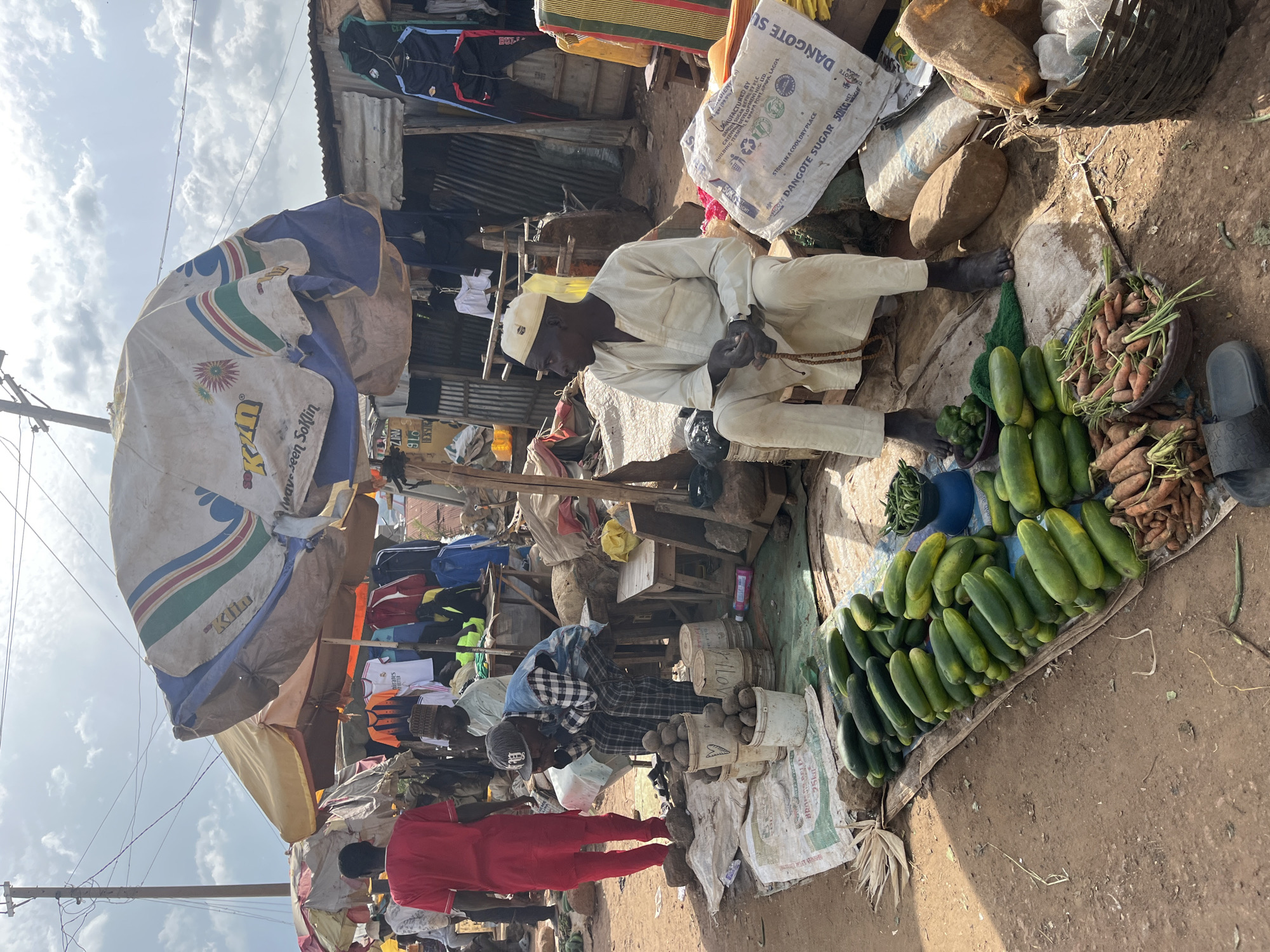 The image depicts an outdoor market scene in Jos, Nigeria, under a cloudy sky. In the foreground, a pile of cucumbers lies on a mat, with a basket of carrots and a bowl of green peppers nearby. To the right, a woven basket overflows with produce. Behind the vegetables, two men are seated on the ground; one wears a white cap and traditional clothing, holding prayer beads, while the other, in a checkered shirt and cap, appears to be resting his head in his hands. Further back, a person in a red outfit stands, facing away from the camera. Several market stalls with colorful umbrellas are visible, displaying clothing items. The ground is a mixture of dirt and scattered debris, characteristic of an open-air market.