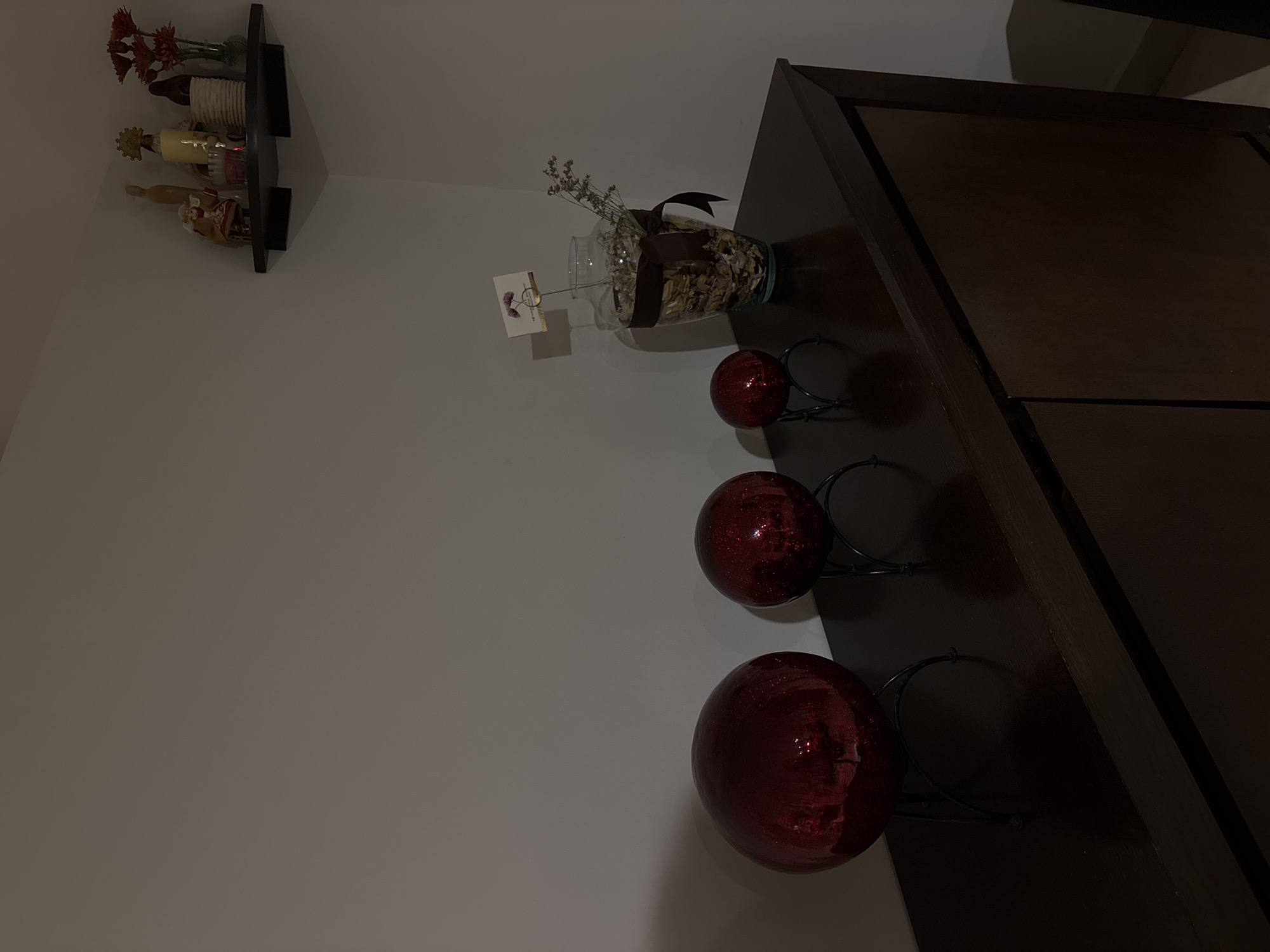 The image shows a low-lit interior scene. A dark brown wooden cabinet stands against a white wall. On top of the cabinet are three large, shiny red spherical decorations held in black metal stands. To the left of the spheres, a clear glass vase containing dried flowers and leaves, tied with a brown ribbon, sits near a small card with writing. Above and to the left of the cabinet, a black shelf holds a variety of decorative objects, including what appear to be figurines and floral arrangements. The location is possibly a residence in Puebla, Mexico, judging by the architectural style and typical decor.