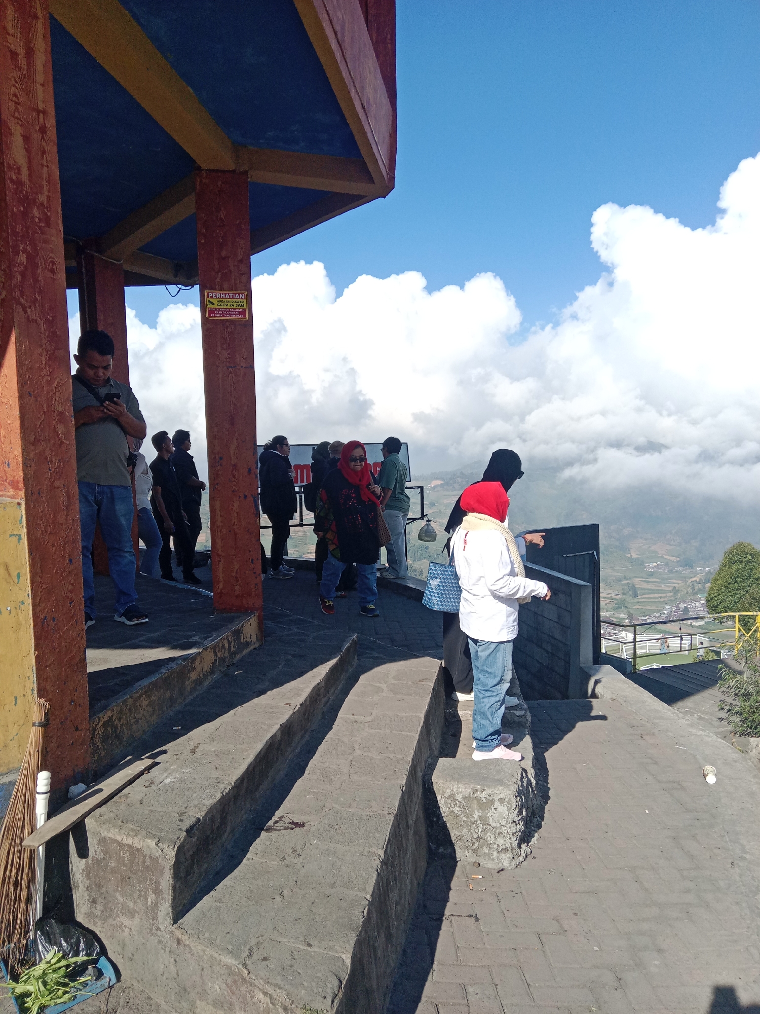 In Tieng, Indonesia, a group of tourists are seen standing under a covered structure, looking out at a scenic view. The structure has red, yellow, and blue elements and is supported by pillars. A sign with Indonesian text is visible on one of the pillars. The tourists, dressed in casual attire, are positioned on steps and a paved walkway overlooking a valley with houses and terraced fields. Some are looking at their phones, while others are observing the landscape, with one person pointing towards the view. The sky is blue with scattered white clouds, suggesting a clear day. The environment appears to be a viewpoint or a rest stop, likely at an elevated location given the panoramic view of the valley below.