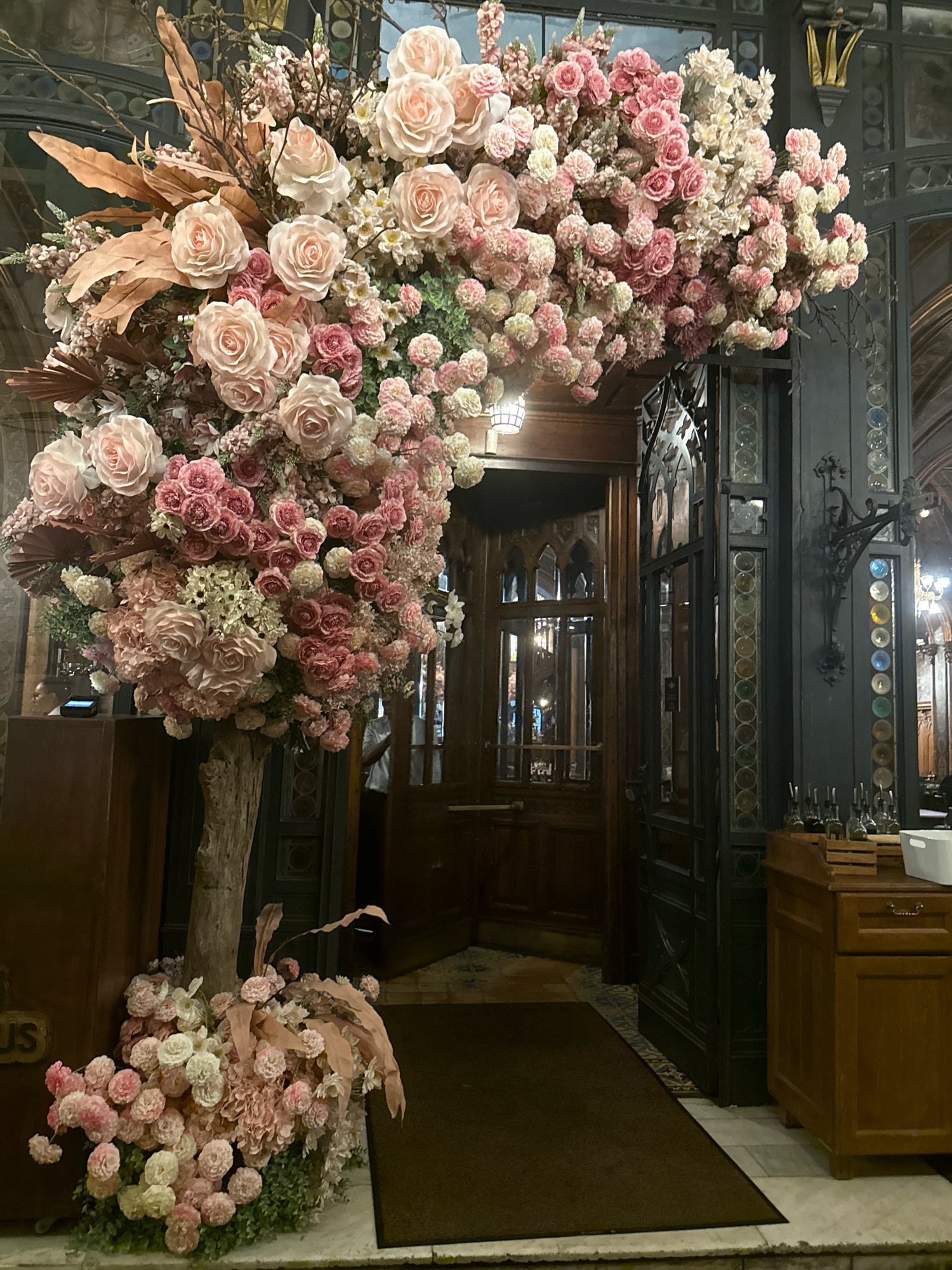 A tall, rustic tree trunk serves as the base for an abundant floral arrangement. The arrangement features a multitude of pink and white roses of various sizes, interspersed with smaller pink flowers, hydrangeas, and dried tan leaves. The floral display extends upwards and outwards, creating a grand arch over a doorway. The doorway itself is made of dark wood with gothic-style glass panels. To the left of the doorway, a dark wooden cabinet with gold lettering is visible. To the right, a dark metal gate with decorative glass panels and a dark wooden counter with bottles on top is present. The floor is tiled, with a brown mat in front of the doorway. The overall impression is one of opulent decoration within an established architectural setting, possibly a restaurant or hotel lobby.