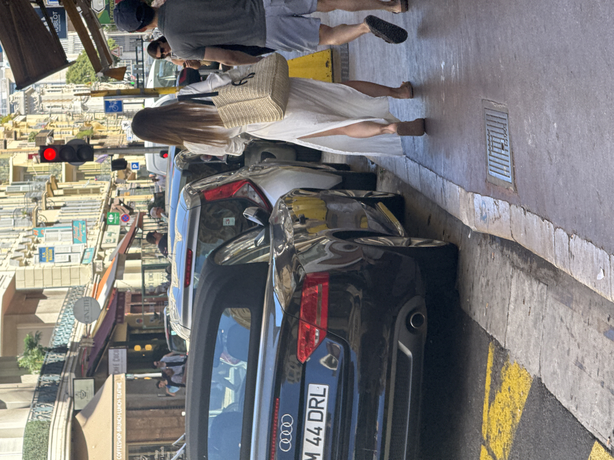 This image depicts a street scene in Nice, France, likely on the Promenade des Anglais or a similar prominent street due to the architecture and the presence of a traffic light. A black Audi convertible is parked on the right side of the street, its license plate reading "M 44 DRL". In front of the Audi, other vehicles are parked, including a white car and a silver car. On the sidewalk to the right of the parked cars, two individuals are walking away from the camera. The person in front is a woman wearing a long, white, slit dress and flat sandals. She is carrying a large straw tote bag over her right shoulder. Behind her, a man in a grey t-shirt and blue shorts is walking. He is wearing dark sandals. Further down the street, more buildings with awnings and signs are visible, including a sign for "HUBOLT". A red traffic light is illuminated, indicating a stop. The street is paved with asphalt, and the sidewalk is made of concrete. Yellow markings are present on the road, suggesting a parking zone. The overall impression is a sunny day in a bustling European city.