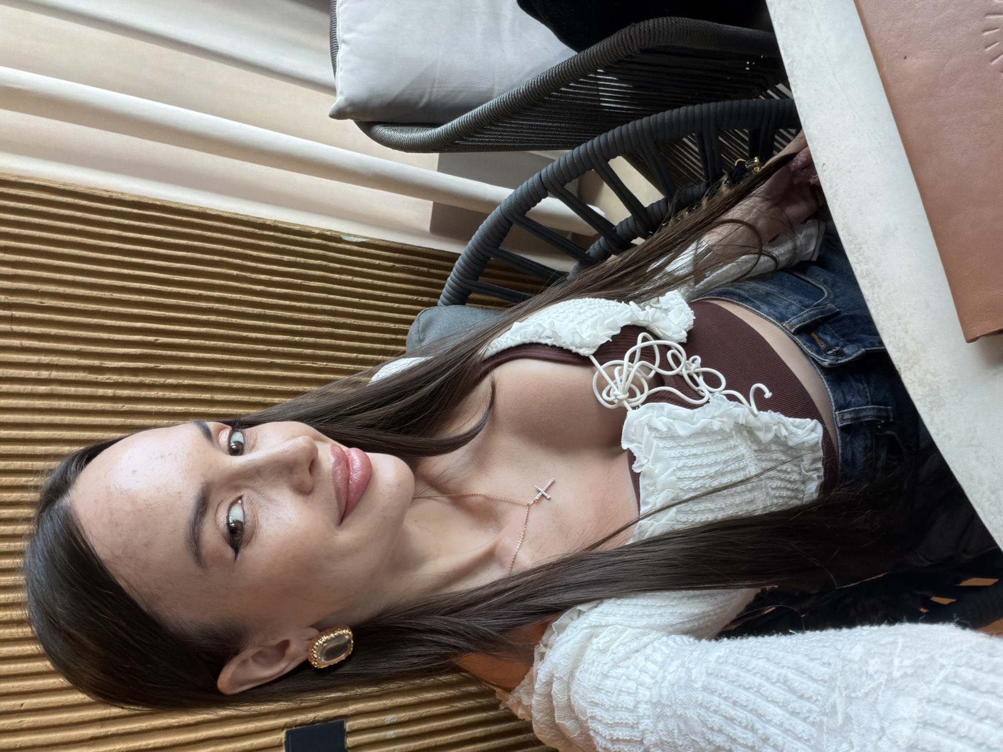 A woman with long, dark hair and light skin is sitting at a table in an establishment with a textured golden wall and beige curtains in the background. She is wearing a white crocheted top with a brown bralette underneath, and blue jeans. A silver cross necklace with small gems is visible around her neck. She is also wearing a large, ornate gold earring on her left ear. Her right hand is resting on the table, and her left hand is obscured by the table. Two dark woven chairs are visible behind her, one with a light grey cushion. The environment appears to be indoors, possibly a restaurant or cafe. This scene is located in Saint Petersburg, Russia.
