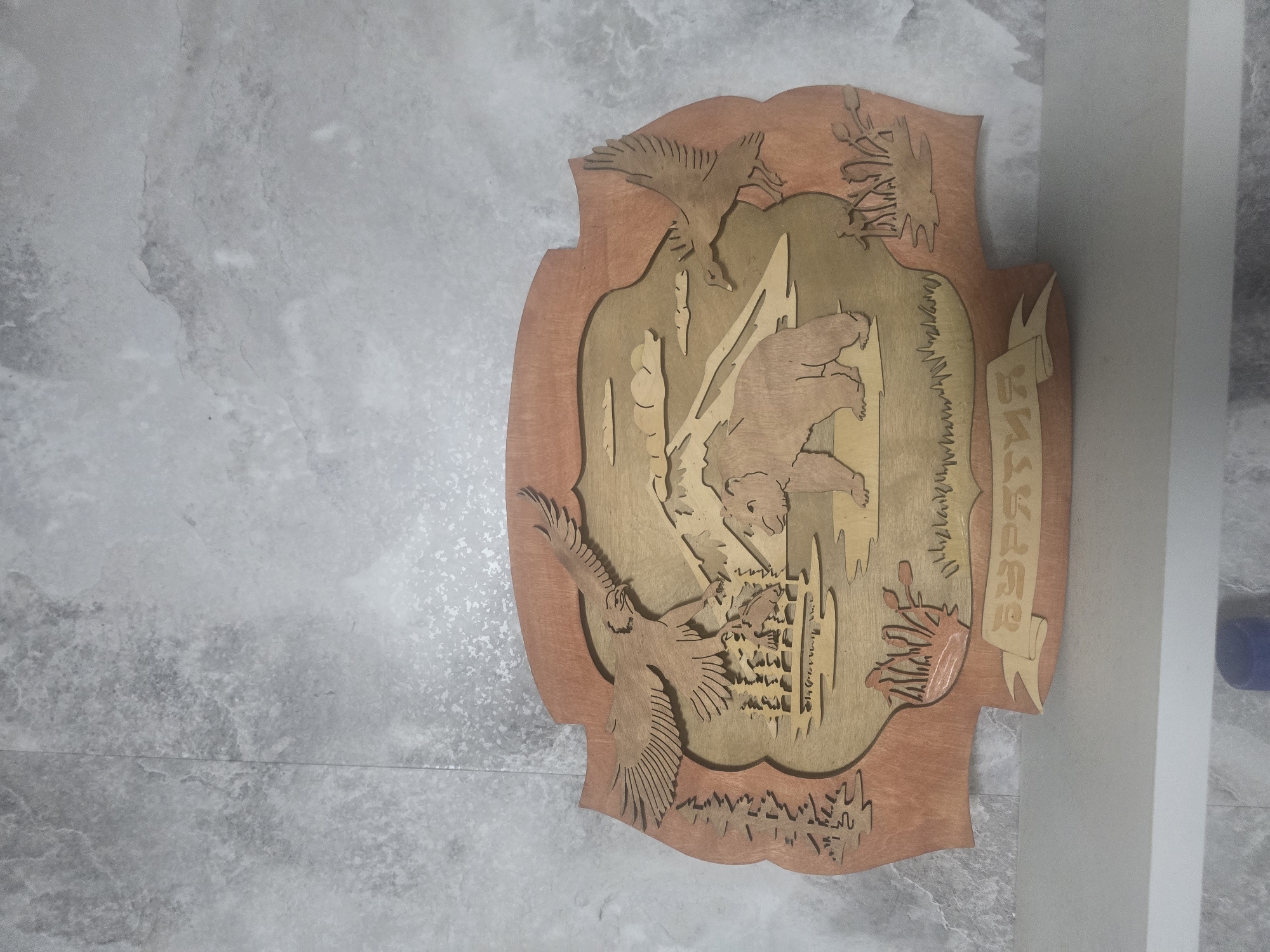 This is a photograph of a wooden relief carving depicting a wilderness scene, with the name "БУРЯТИЯ" (Buryatia) inscribed on a banner at the bottom. The carving is mounted on a wall with a textured gray finish.
The carving itself features a brown bear standing near a body of water, with mountains in the background. On the left, an eagle is shown swooping down towards the water, carrying a fish in its talons. Another bird, possibly a duck, is flying on the right side of the carving. The foreground includes representations of trees and vegetation.
The overall context of the image, as suggested by the inscription "БУРЯТИЯ", points to a cultural or geographical representation related to the Republic of Buryatia, a federal subject of Russia located in southeastern Siberia. Given the image depicts a wilderness scene, it is plausible that this carving is displayed in a location within or associated with the city of Maksimiha, Russia, which is situated on the eastern shore of Lake Baikal, a region known for its natural beauty and wildlife. The carving is well-lit, allowing for clear observation of its details.