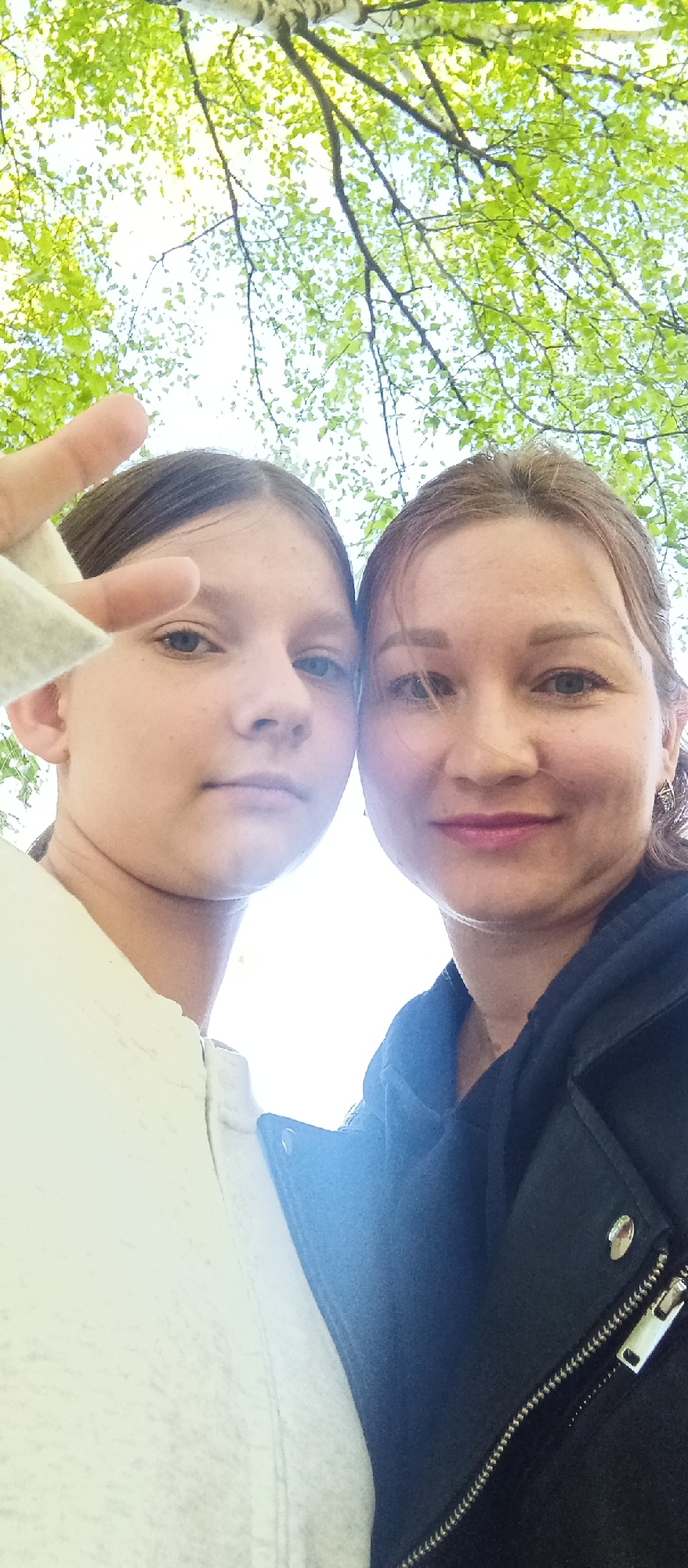 The image captures two individuals, a younger person and an adult female, standing close together and facing the camera in a selfie-style shot. They appear to be outdoors, as the background shows trees with green leaves against a bright sky. The younger person, positioned on the left, is making a hand gesture, possibly a peace sign or similar. They are wearing a light-colored garment. The adult female, to the right, has a dark jacket with a zipper and silver buttons visible. The location appears to be a natural setting, possibly a park or wooded area, and is located in the city of Unknown, Unknown.