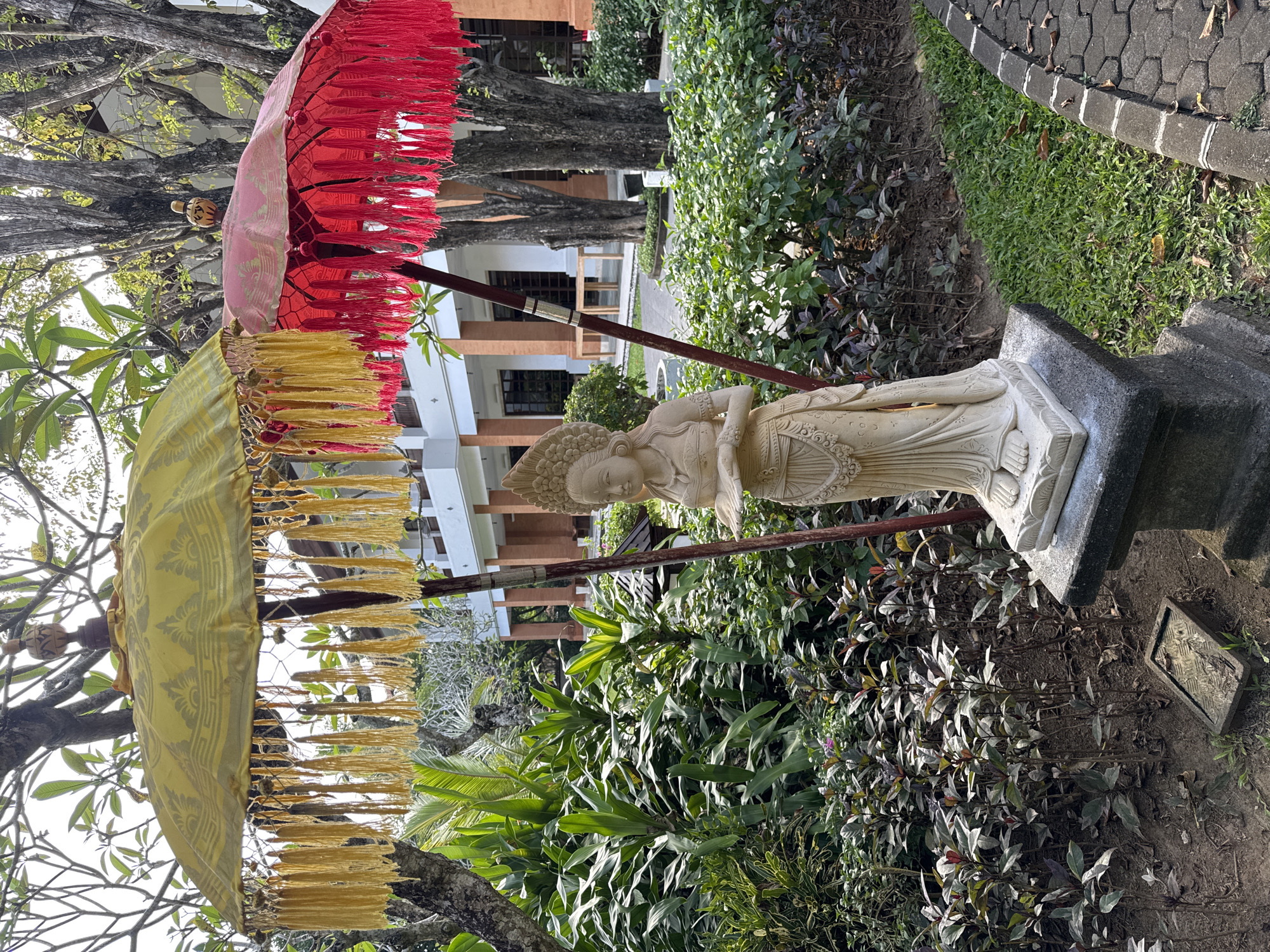 The image depicts a stone statue of a deity in a garden setting in Kuta Selatan, Indonesia. The statue is light in color and stands on a dark grey pedestal. Overhead, two Balinese parasols provide shade. One parasol is yellow with gold fringe and the other is red with red fringe. They are supported by a dark wooden pole. Lush tropical vegetation surrounds the statue and umbrellas. In the background, a two-story building is visible. A cobblestone walkway borders the garden.