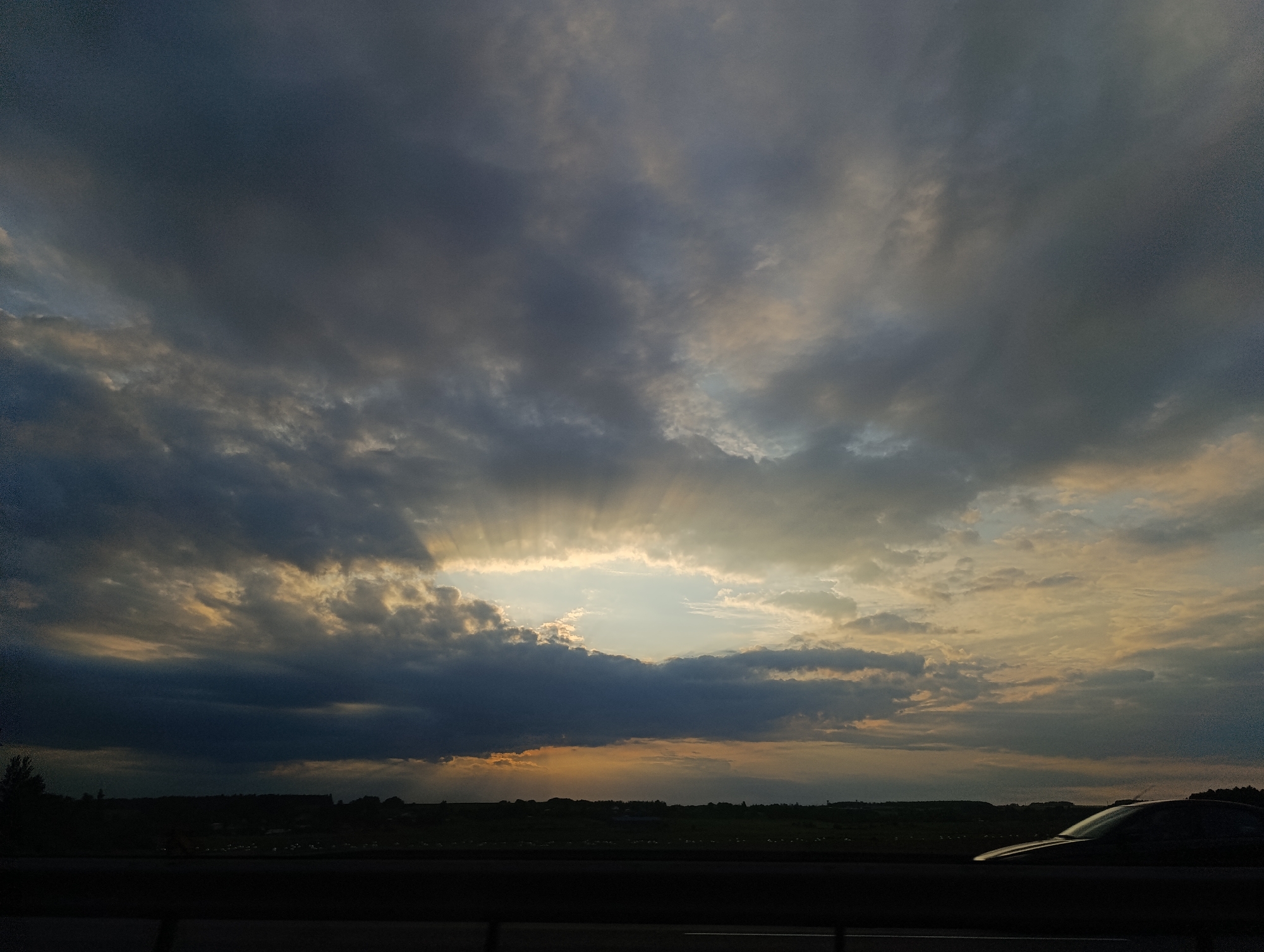 The image depicts a dramatic sunset with crepuscular rays filtering through a sky filled with dark, stormy clouds. A sliver of clear sky with lighter clouds and the setting sun is visible in the center. Below the sky, a dark silhouette of a landscape stretches across the horizon, featuring trees, buildings, and fields. In the foreground, a dark horizontal barrier runs across the bottom of the frame, and a car is partially visible on the right, its curved roof reflecting the dim light. The scene appears to be captured from a moving vehicle on a road, suggesting a highway or a similar thoroughfare. The overall mood is atmospheric and somewhat ominous due to the interplay of light and shadow.