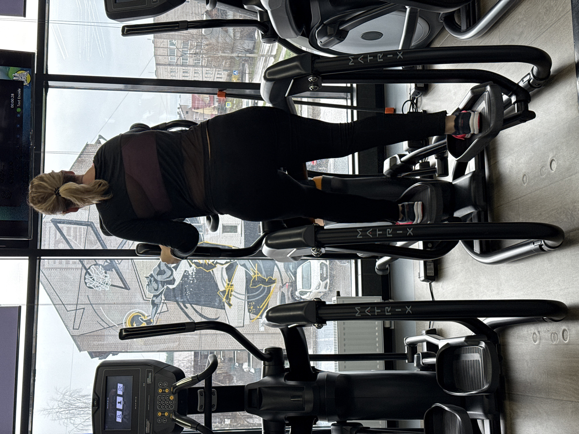 A woman with blonde hair in a ponytail is working out on an elliptical machine in a gym. The machine is black and has the brand name "MATRIX" on it. The woman is wearing black leggings, a black long-sleeved shirt with a maroon underlayer, and sneakers with pink accents. She is facing a large window with a view of a city street. Outside the window, there is a mural on a building depicting a basketball player and a basketball hoop. The mural features yellow and black paint. The buildings outside the window include several large, drab apartment buildings with visible windows. The weather outside is overcast and the street appears wet. There is a television screen on the left side of the frame. The gym has a gray floor. The city is identified as Votkinsk, Russia.