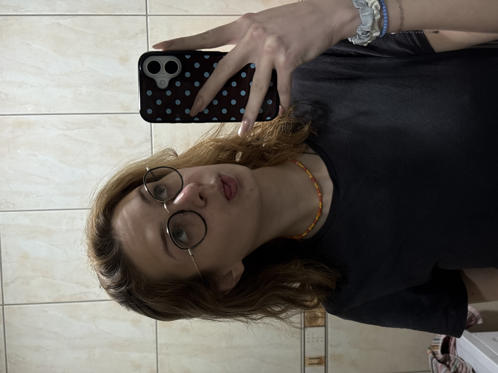 A person is depicted taking a mirror selfie indoors. The image is rotated 90 degrees clockwise, showing the individual's head on the lower left, facing upwards. The person has light brown or blonde wavy hair and is wearing round-framed glasses. They are making a playful expression, sticking their tongue out slightly.
The individual is wearing a plain black t-shirt and a colorful beaded necklace with red, orange, and yellow beads around their neck. On their right wrist, multiple accessories are visible: a light blue scrunchie, a blue beaded bracelet, and a delicate silver-colored chain or bracelet.
Their right hand is holding a black smartphone with a light blue polka dot case, which is visible in the reflection, showing the phone's dual camera module. The person's fingers have neatly shaped nails, possibly with a clear or light polish.
The background consists of light-colored, possibly beige or off-white, rectangular tiles with visible grout lines, suggesting a bathroom or a similar tiled indoor space. In the bottom right corner of the original image, a small portion of what appears to be a washing machine or laundry appliance, with some striped fabric draped over it, can be seen.
The lighting appears to be artificial indoor light. There is no visible text in the image. While the location context of Kragujevac, Serbia, was provided, there are no specific visual cues within the image itself to confirm this geographic location.