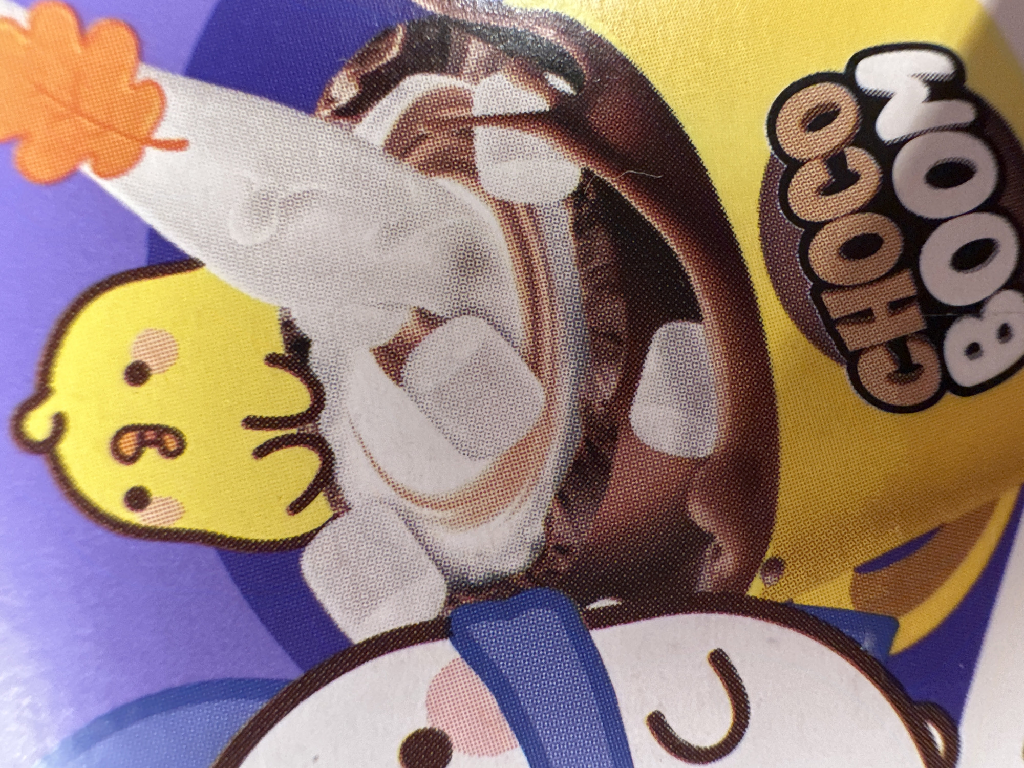The image is a close-up of a product wrapper, likely for a snack or confection. The dominant colors are purple, yellow, and brown, with accents of white and orange.
The main subjects are cartoon characters. On the left, a yellow, chick-like character with a simple face, including dots for eyes and a stylized mouth, is visible. To the right of the chick, a large, brown, swirled object that resembles a chocolate dessert or swirl is prominently featured, with white dollops that could be cream or marshmallows embedded within it. Part of a white, cone-shaped object with an orange leaf shape at its tip is also visible, extending from behind the chocolate swirl. At the bottom of the image, another cartoon character, mostly white with round pink cheeks and dark eyes, is peeking out. This character appears to have a blue ribbon or bow around its neck.
The text "CHOCO BOOM" is emblazoned on the yellow part of the wrapper in a bold, rounded font with a dark outline and a brown, textured fill.
There are no clear indications of time of day, weather, or a specific real-world location, other than the provided context of Minsk, Belarus. The overall aesthetic suggests a playful and appealing design, common for food products aimed at a broad audience. The scene is static, depicting an illustration on a package. The emotions conveyed are likely cheerful and fun due to the cartoon characters and bright colors.