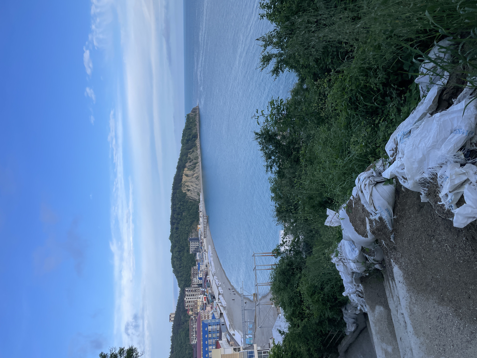 View of the Black Sea and the beach of Olginka, Russia. The view is captured from an elevated position with a concrete wall and sandbags visible in the foreground, along with lush green bushes. Beyond the wall, the coastline stretches out, revealing a sandy beach dotted with white beach umbrellas and buildings. Further out, the Black Sea extends into the distance, with a long, narrow peninsula jutting into the water, covered in trees. The sky above is partly cloudy.