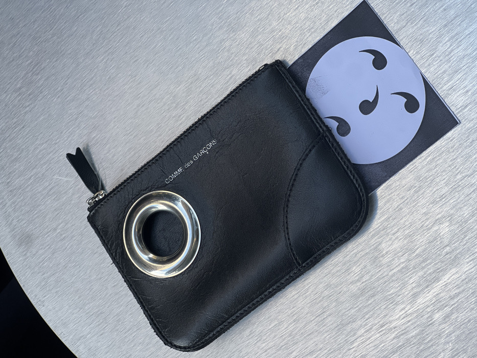 The image displays a black leather Comme des Garçons pouch or wallet positioned on a brushed metallic surface, likely a table, exhibiting horizontal graining. The pouch is oriented horizontally and features a prominent, polished silver-colored circular grommet on its upper left portion. A zipper closure runs along the top edge, equipped with a small, black leather pull tab. The brand name "COMME des GARÇONS" is visible in light-colored text on the upper right side of the pouch. Beneath the lower right edge of the pouch, a black card or paper is partially visible, showing a white circular graphic containing three black, comma-shaped or tomoe-like symbols. The background beyond the metallic surface is dark and indistinct. No individuals, actions, or interactions are present in the scene. The image contains no visual cues to ascertain a specific location within Tel-Aviv, Israel.
