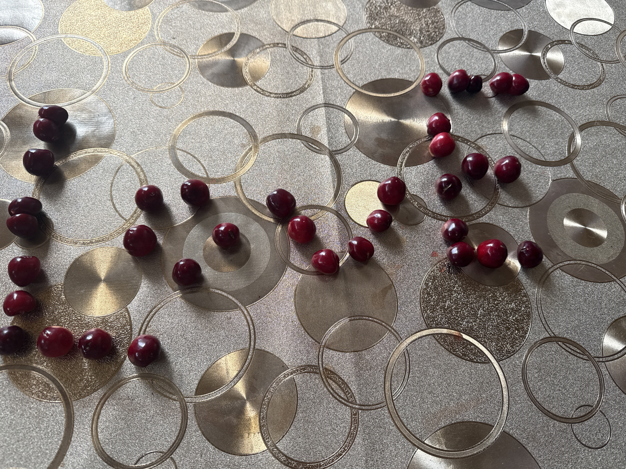 The image shows a pile of red cherries scattered on a patterned surface. The surface has a metallic sheen and features a repeating pattern of concentric circles in various shades of gold and silver. The cherries are dark red and appear to be fresh. Some are whole, while others show slight indentations. The lighting suggests an indoor setting, casting subtle shadows. There are no people or any other discernible objects in the scene. The image's composition is from a top-down perspective.