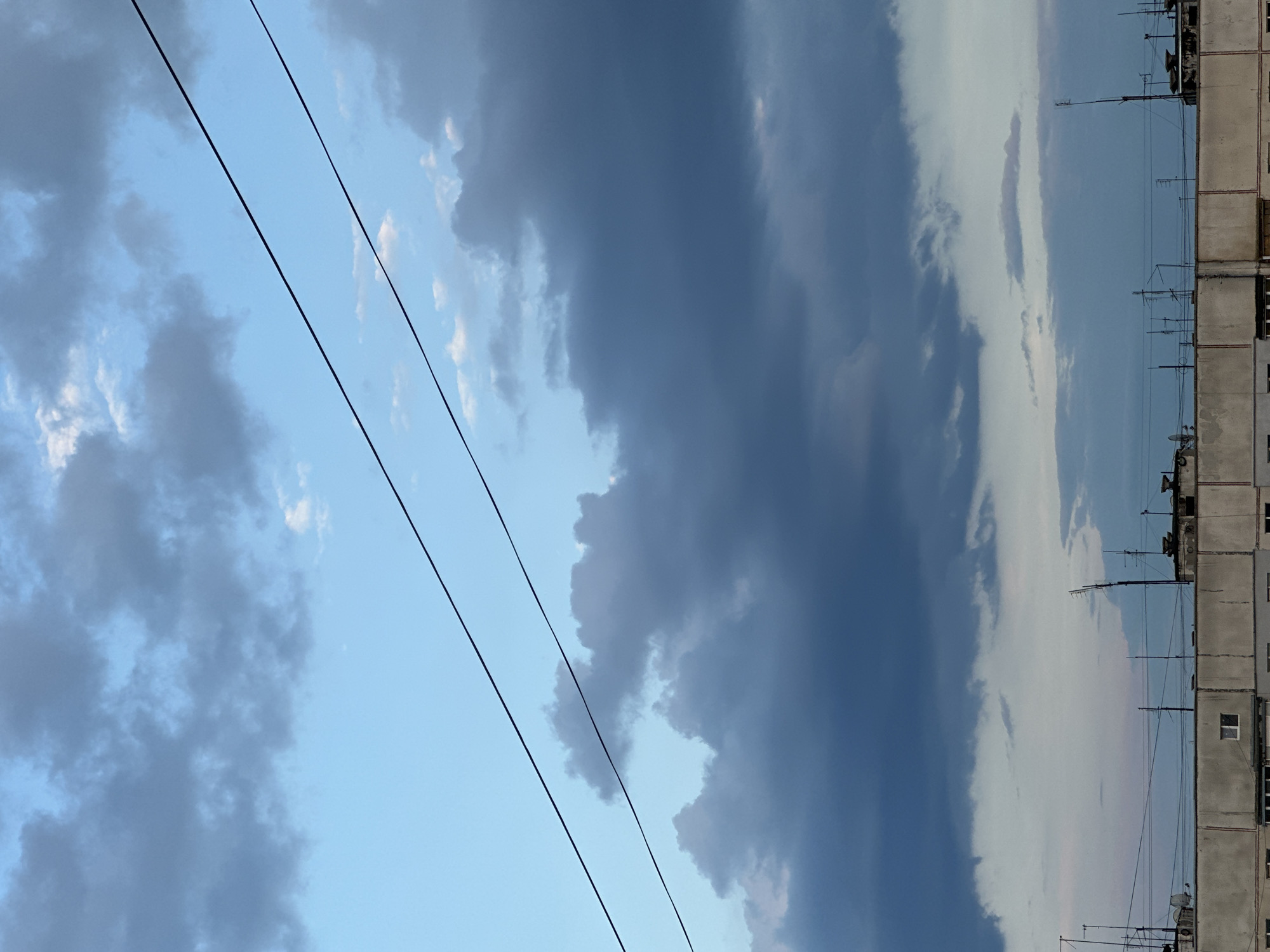 A view from a rooftop in an urban setting, looking up at a sky filled with clouds and crisscrossed by power lines. The sky transitions from a pale blue at the top to darker, more ominous clouds in the middle and lower sections. The clouds are a mix of grays and blues, with some lighter patches. Two dark power lines stretch diagonally across the upper portion of the image. Below the sky, the top of a building is visible, featuring a flat roof with various antennas and satellite dishes. The building's facade is light gray and appears to be constructed from concrete. The overall impression is one of an overcast or approaching storm in a city.