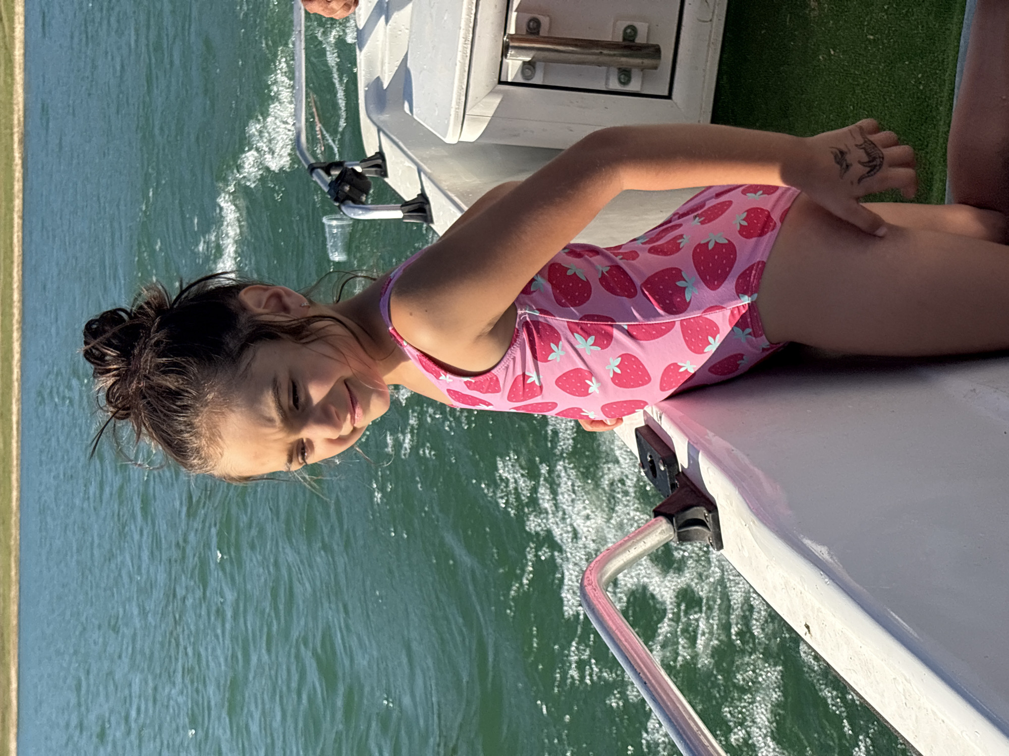 A young girl with her dark hair pulled up in a messy bun stands on a boat. She is wearing a pink swimsuit with a strawberry pattern and is looking to her left with a slight smile. The boat is on a body of water, likely a lake, with a shoreline visible in the background. The water is a vibrant turquoise and appears to be moving, creating small waves and foam. The boat has white siding and a silver railing. The overall scene suggests a recreational activity on a sunny day. The location is identified as Golyama Zhelyazna, Bulgaria.