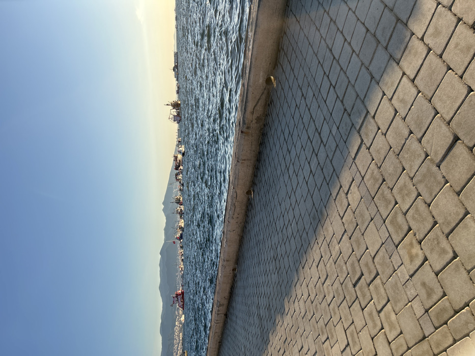 The image shows a cobblestone walkway along the waterfront in İzmir, Türkiye. The walkway is located near the coordinates 38.438 latitude and 27.141 longitude. In the distance, there are numerous boats and ships. The water is a calm blue, and the sky is a clear blue. The sun is setting, casting a long shadow over the walkway. There are no people visible in the image.
