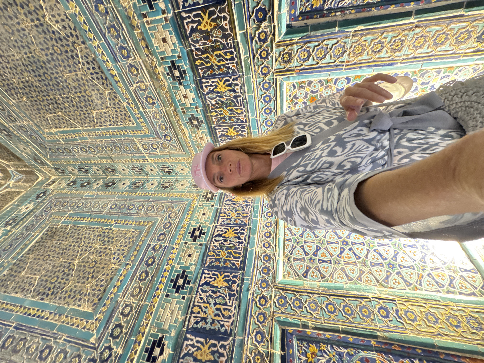 The image is a selfie of a fair-skinned woman with blonde hair, wearing a pink cap and sunglasses, standing in front of a highly decorated, tiled ceiling. She is wearing a patterned grey and white top with a grey ribbon. The ceiling is predominantly blue, turquoise, and yellow, featuring intricate geometric and floral patterns.
The image appears to be taken at a historical or religious site known for its elaborate tilework. Based on the color palette, pattern style, and city provided, the location is likely within Samarkand City, Uzbekistan. The tilework is characteristic of Islamic art and architecture common in Central Asia.