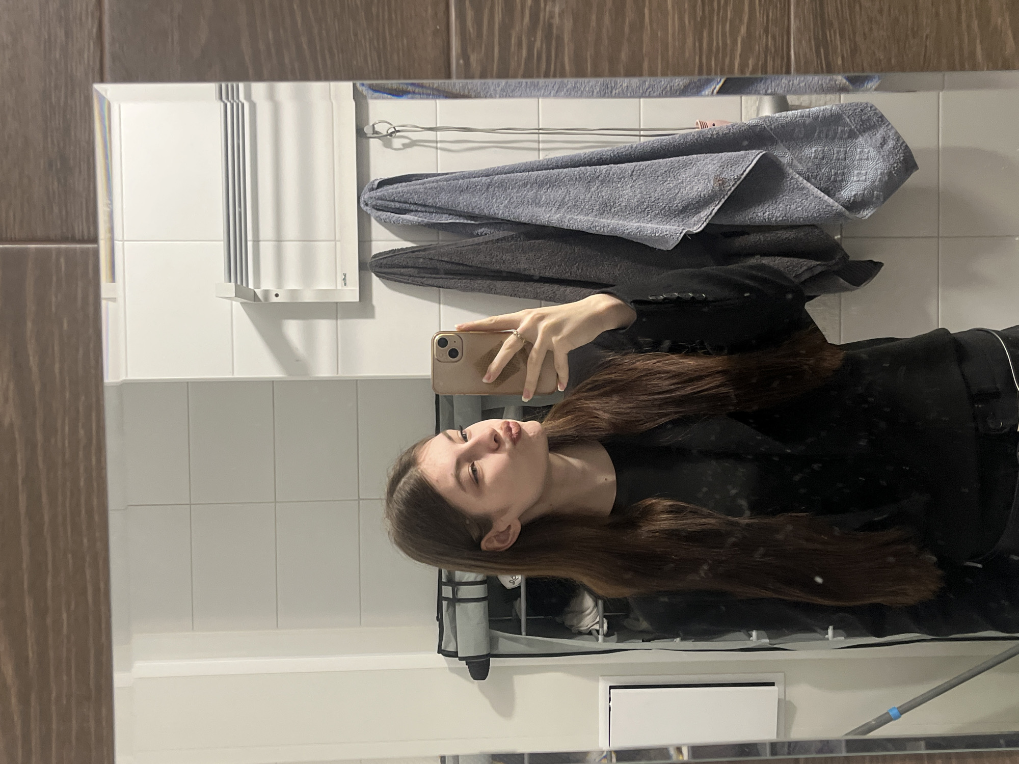 A young woman is taking a selfie in a bathroom mirror. The bathroom has white tile walls and brown wood-look tile on the left. A towel rack with folded gray and blue towels hangs on the wall above a white cabinet. The woman is wearing a black top and holding a tan-colored smartphone. She has long brown hair and is making a kissing face. No information in the image suggests it is located in Saint Petersburg, Russia.