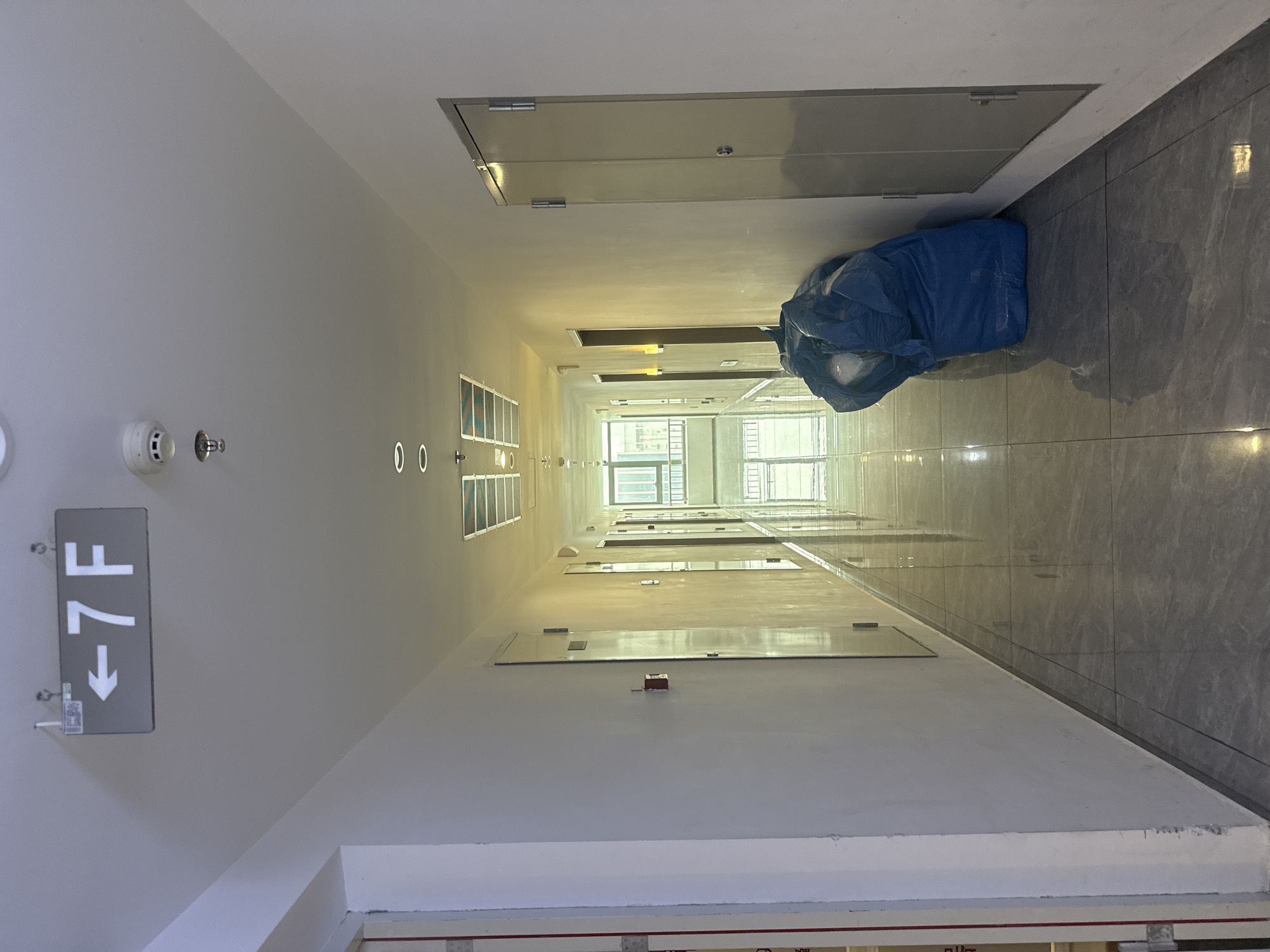 The image shows a long, well-lit hallway in a building, likely a hotel or apartment complex. The hallway has white walls and a gray tiled floor that reflects the light. There are multiple doors on both sides of the hallway. On the ceiling, there is a sign indicating "<-7F", suggesting this is the seventh floor. Other ceiling fixtures include lights and a fire alarm system with a sprinkler head. Towards the right side of the hallway, there is a large blue tarp-covered object, possibly garbage or stored materials. The hallway leads to a more open area at the end, where natural light suggests an outdoor or atrium-like space. Based on the available information and the context provided, the location is within the Huangpu District of China.