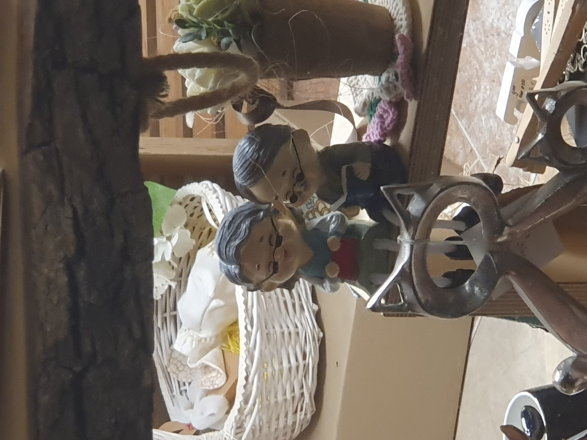 The image depicts a still life arrangement on what appears to be a shelf. Featured are two figurines of elderly people, arranged as if they are kissing, positioned near a white wicker basket containing white fabric and yellow material. Next to them is a decorative metal chandelier. Other decorative elements include a potted succulent and various small crafting items. The location and city (Plovdiv, Bulgaria) cannot be determined from the visual information provided.
