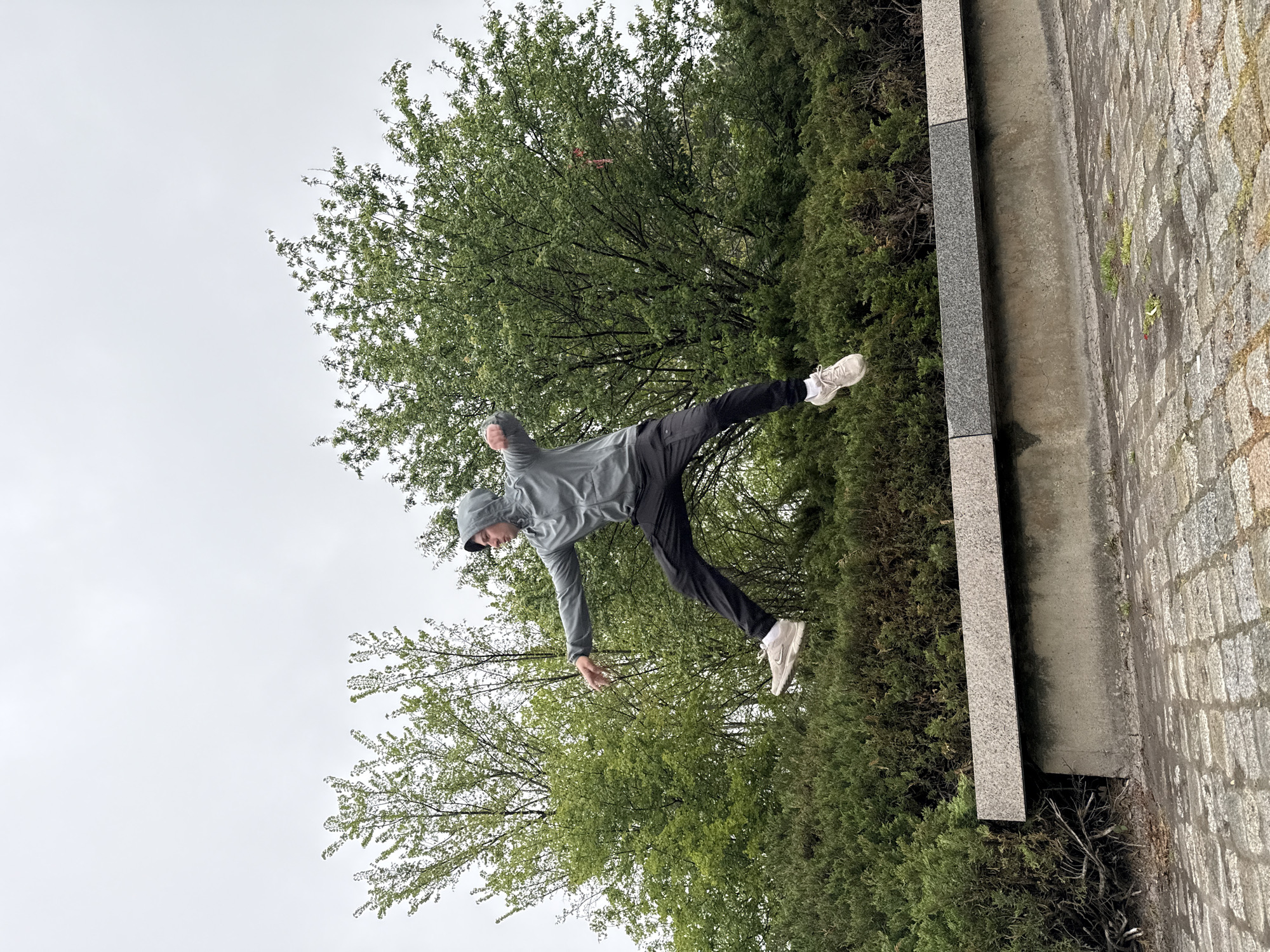 A person wearing a gray hooded jacket, black pants, and white shoes is captured mid-air in front of lush green foliage, possibly trees and bushes. The background includes a gray, overcast sky. Adjacent to the foliage is a gray stone structure, with a paved surface visible in the lower right. The image suggests an outdoor urban setting. The location, according to the request, is in the city of Shumen, Bulgaria, though this is not directly discernible from the image alone. The person appears to be performing an action, possibly jumping or posing.