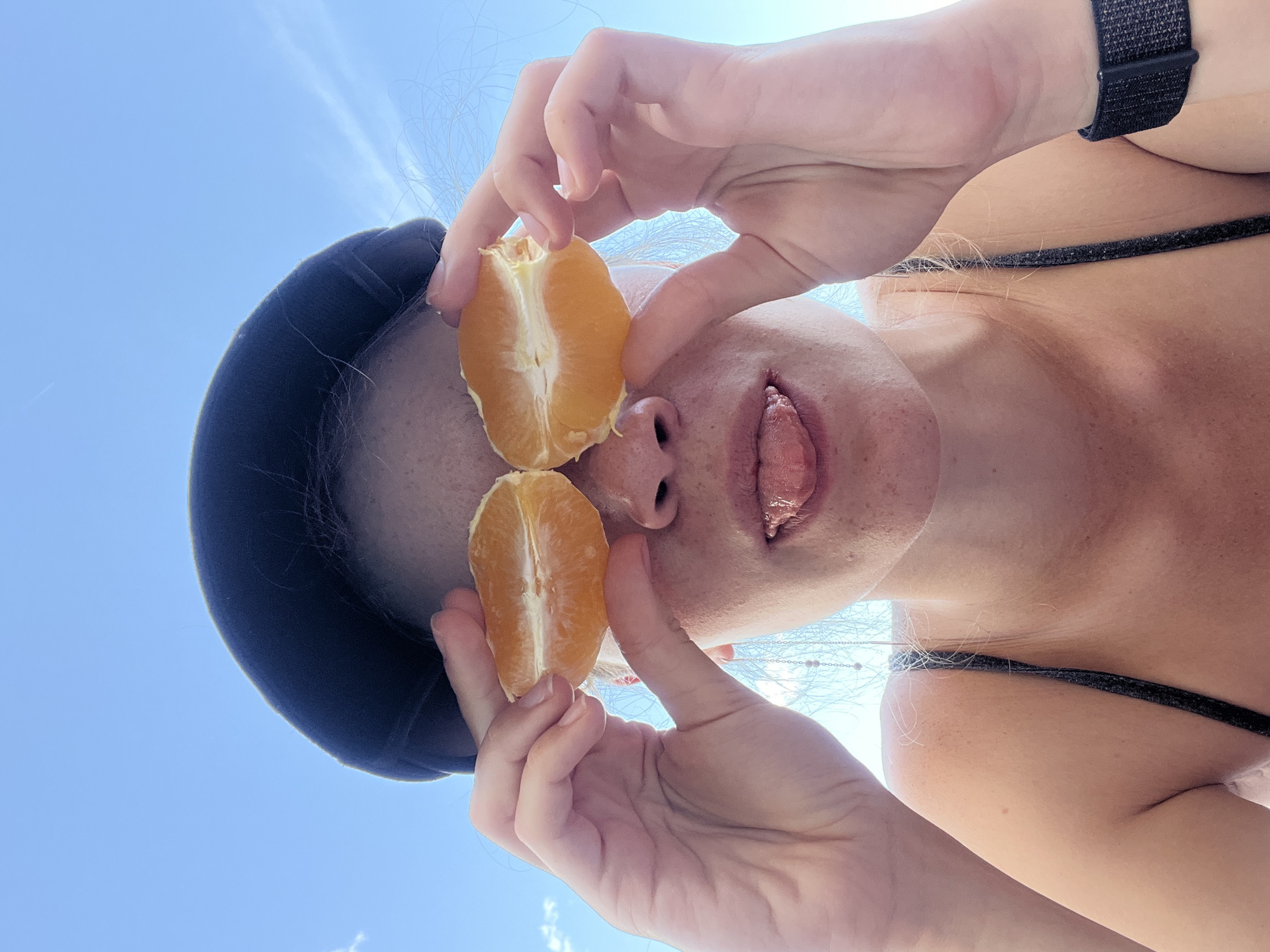 A low-angle shot depicts a person holding two orange halves near their face. They are wearing a black hat, and a black strapped watch is visible on their wrist. The person is wearing a black-strapped top. The background is a clear, light blue sky. The location is unknown; there is no information within the image to determine if it is located in Budva, Montenegro.