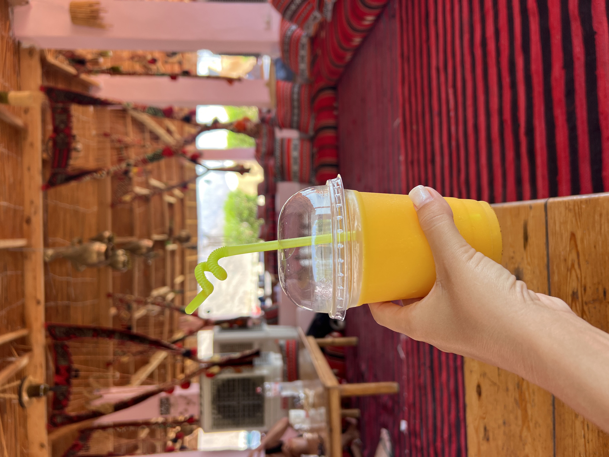 A person's hand holds a clear plastic cup filled with orange juice and a bright green, curly straw in the foreground. The cup is on a wooden table. In the background, there is a seating area with low-lying cushions and pillows, all covered in red and black striped fabric. The ceiling is made of wooden beams and decorated with hanging tapestries and other adornments. A large white air conditioning unit is visible on the left side of the image. The location appears to be an outdoor shaded cafe or seating area, likely in a tourist area within the city of Nuweiba, Egypt, given the traditional decor.