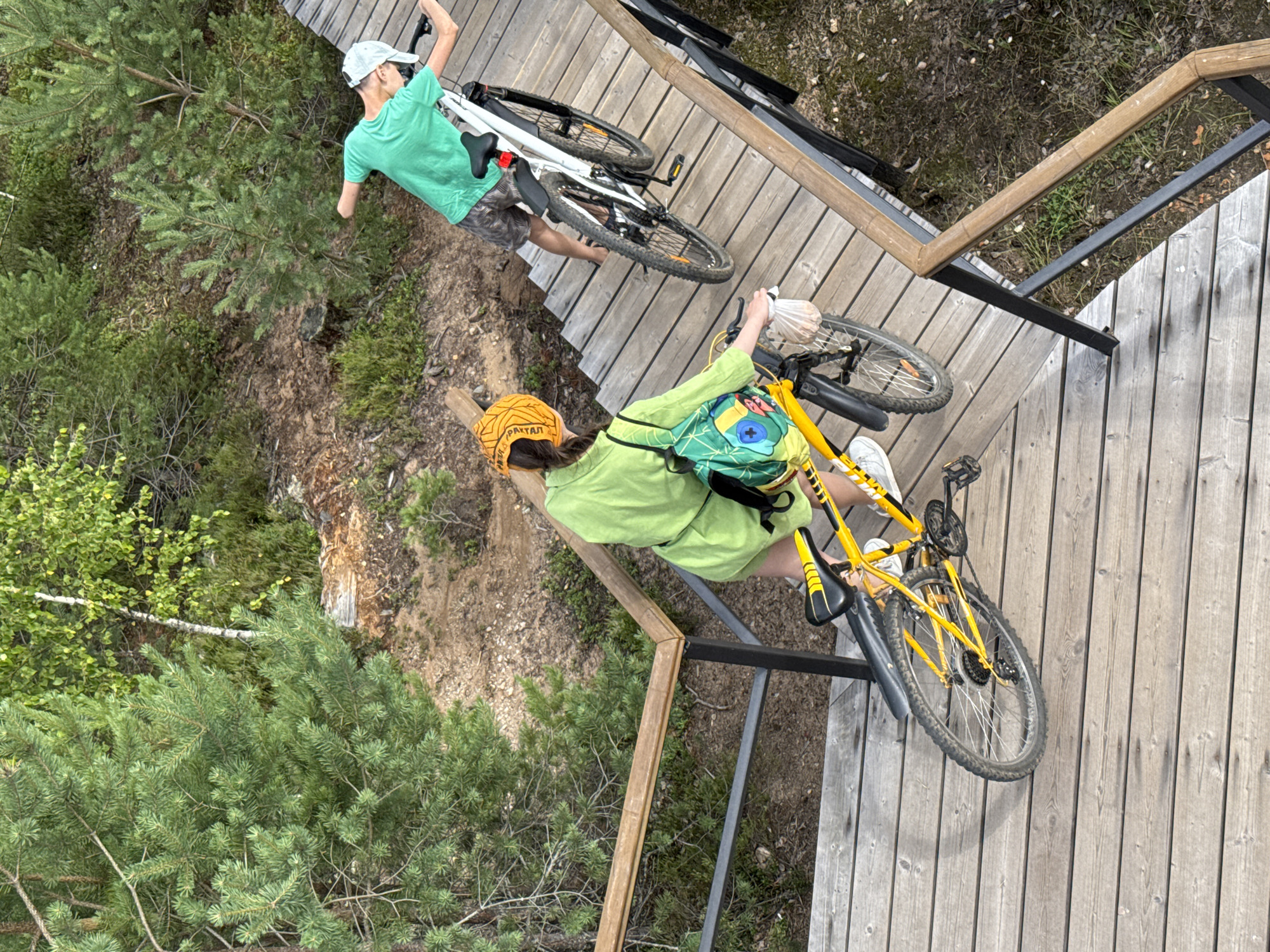 Two people with bicycles are on a wooden walkway in Peski, Russia. The person in the foreground is wearing a lime green outfit and a yellow headband, pushing a yellow bicycle. The person in the background is wearing a green t-shirt and patterned shorts, holding a white bicycle. The walkway is elevated and surrounded by trees and vegetation. The scene appears to be a park or recreational area.