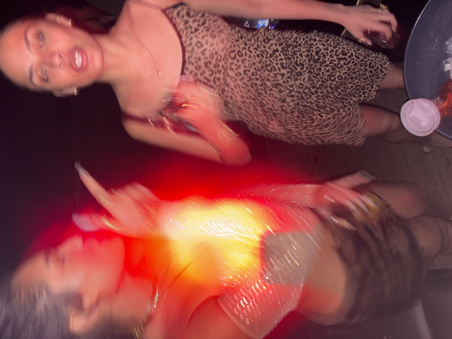 Two women are captured in a blurry, rotated image. The top figure wears a leopard-print dress with thin straps, holding a glass, and has a necklace. The bottom figure, partially obscured by red light, also wears an animal-print outfit, possibly a dress or top with a shiny element. A table holding a drink with ice and a lidded cup is visible in the corner. The overall composition suggests a lively, perhaps dimly lit, social setting.