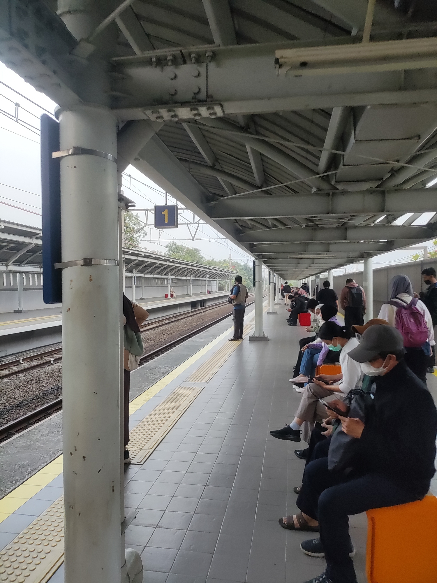 This image depicts a train station platform in Telagamurni, Indonesia, under an overcast sky. Several individuals are seated on orange benches, waiting for a train. Most people are looking at their mobile phones. The platform is covered by a metal roof supported by pillars, and the train tracks run alongside. A sign with the number "1" is visible on the platform. The environment appears clean and orderly, with the train tracks and platform clearly demarcated.